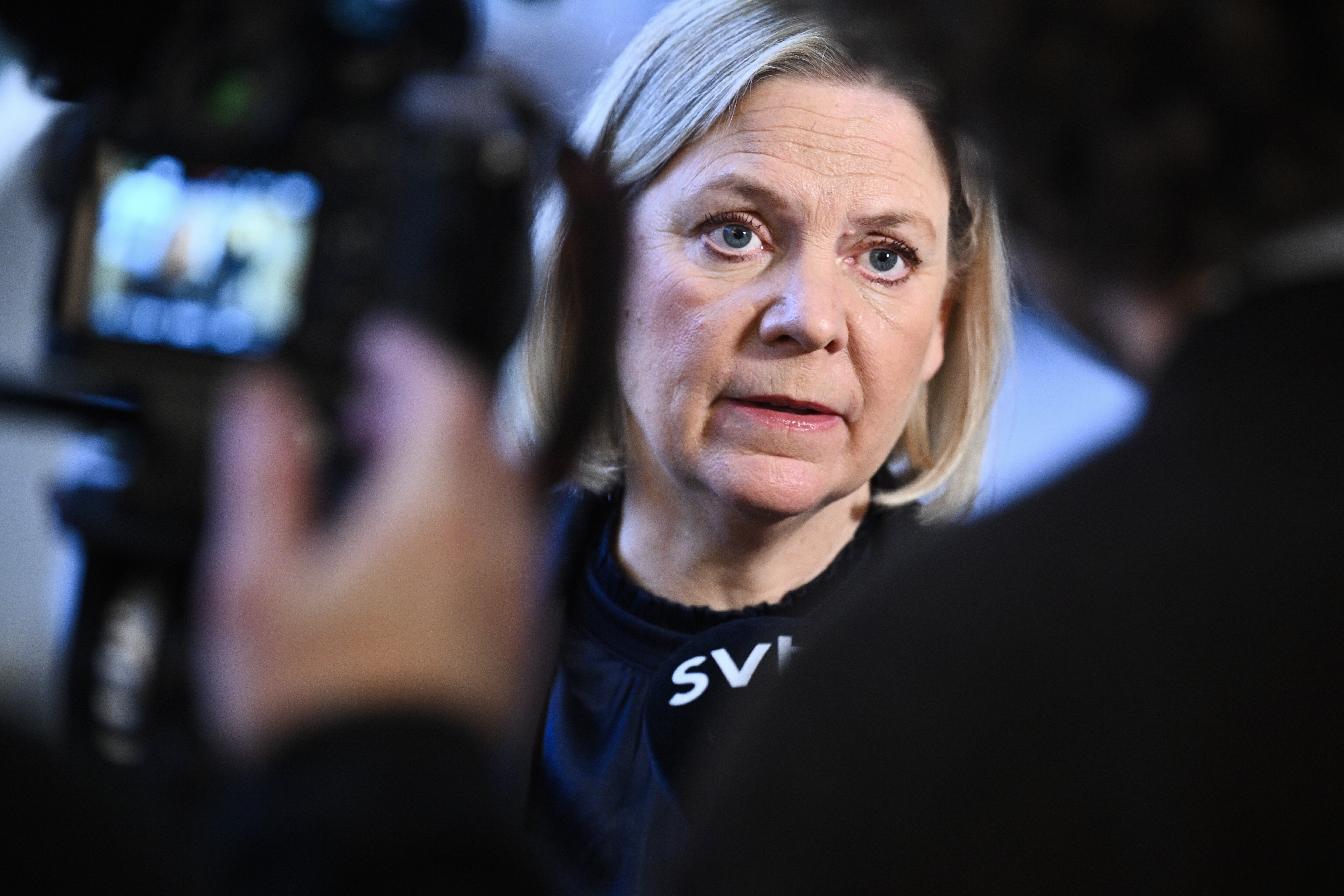 En kvinna med blont hår talar under en intervju, vänd mot kameran, med en mikrofon och kamerautrustning i förgrunden.