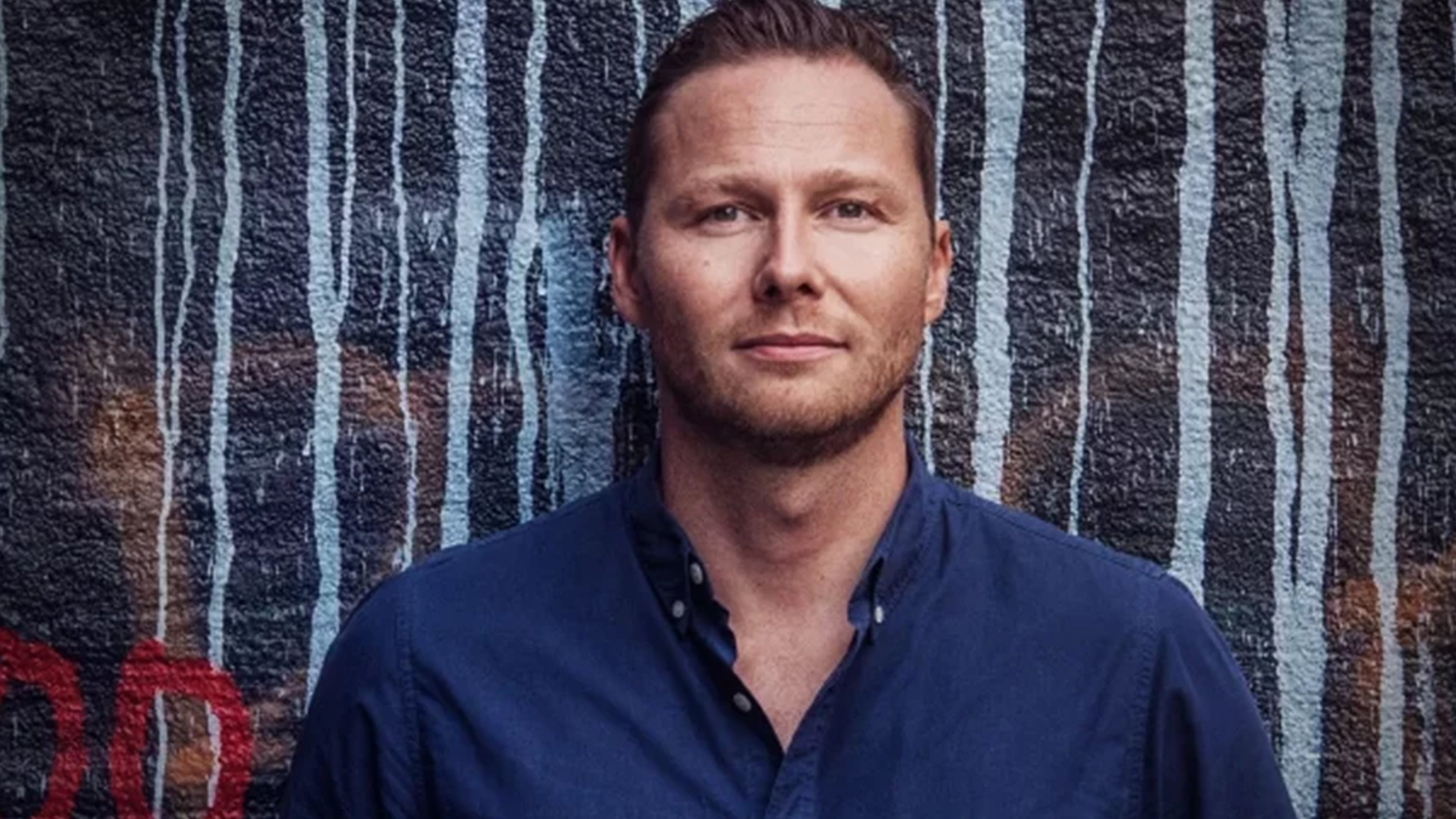 En man med ljust brunt hår och en mörkblå skjorta står framför en målad tegelvägg med vertikala ränder.