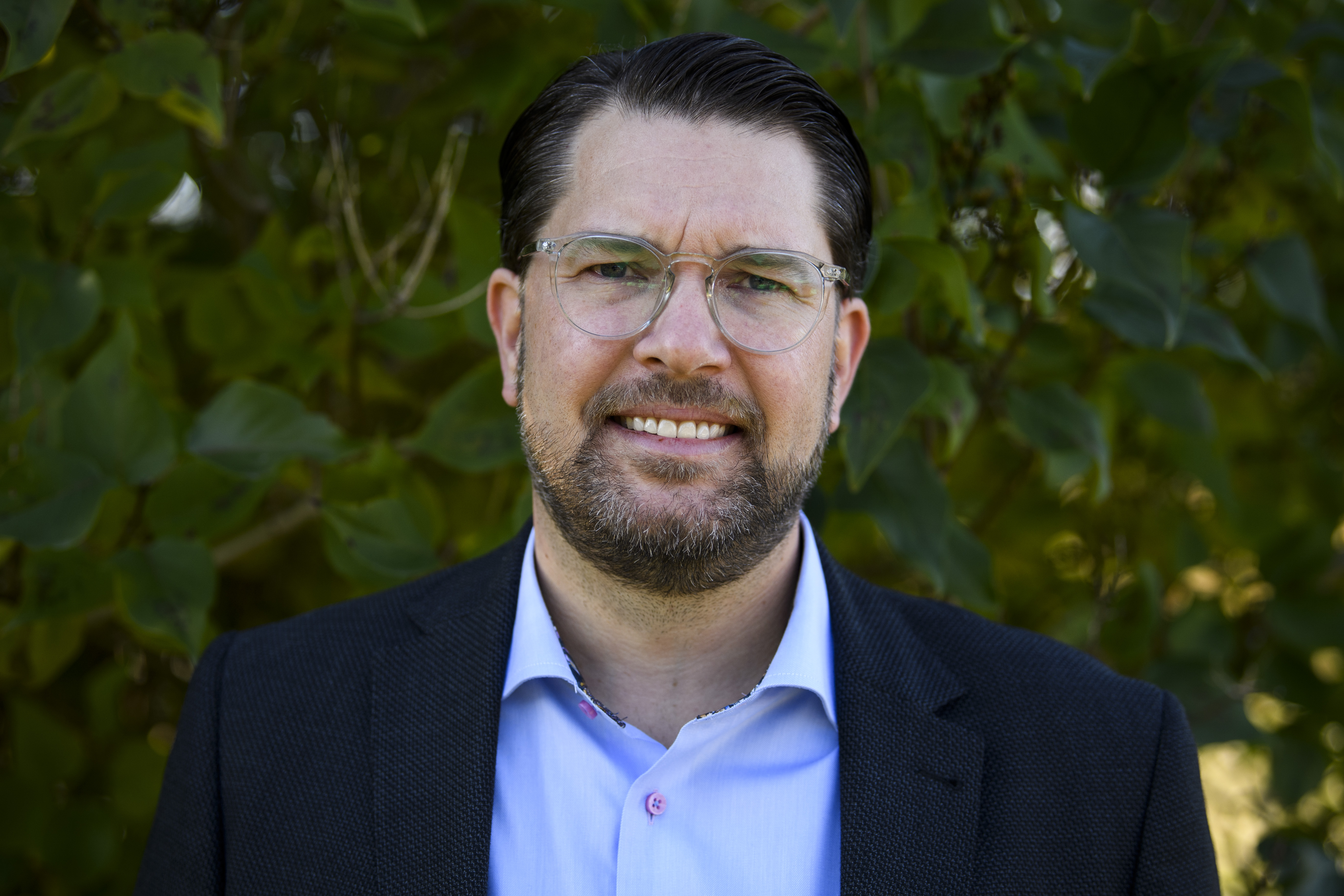 Jimmie Åkesson med kort mörkt hår, glasögon och skägg har en mörk jacka och ljusblå skjorta, står utomhus framför gröna växter.