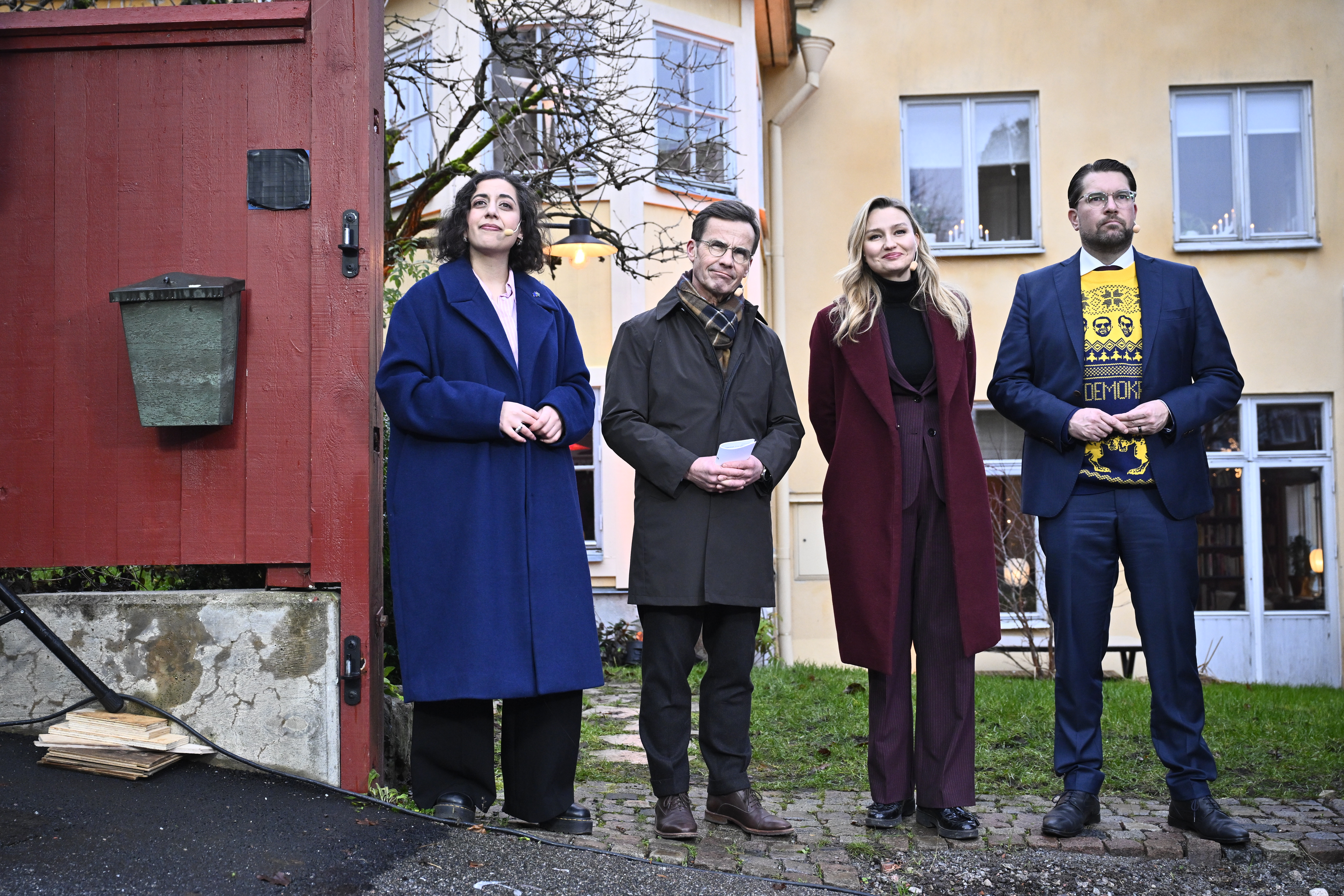 Fyra personer står utomhus framför en röd grind och gula byggnader och poserar för ett foto en mulen dag.