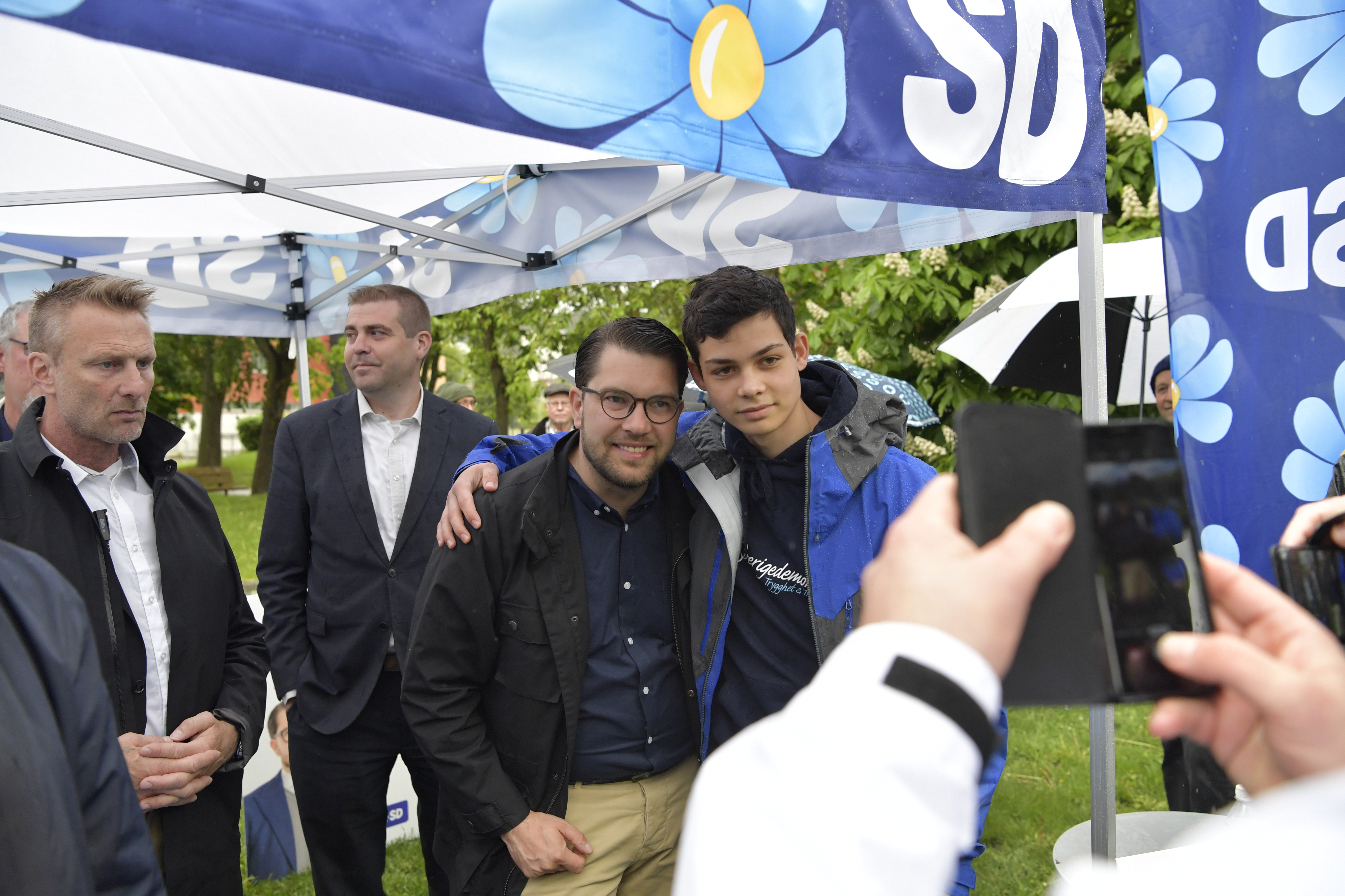En grupp personer poserar för ett foto under ett tält med SD-partiets banderoller, medan någon tar en bild med mobiltelefon.