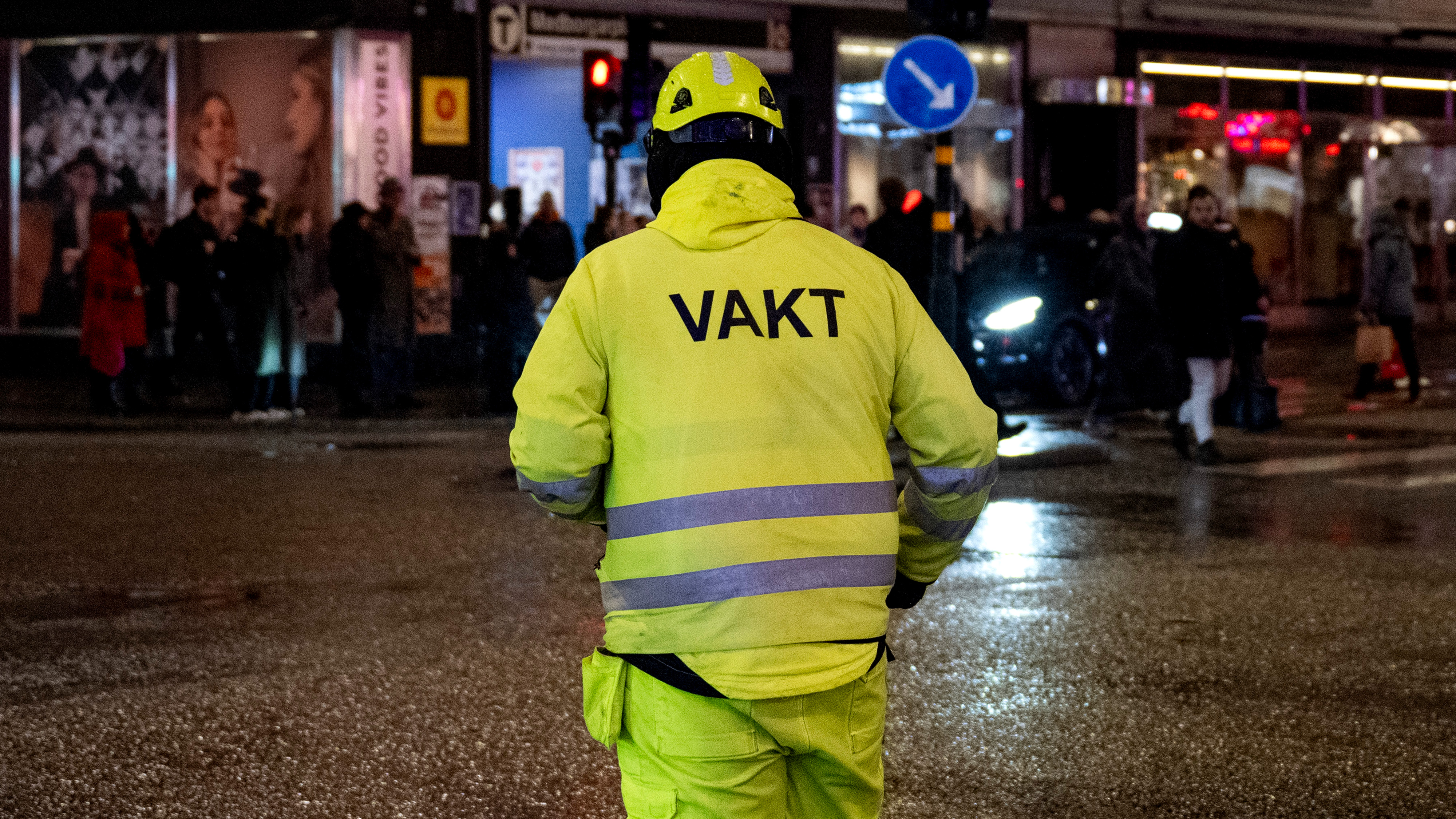 En person med en gul reflexväst märkt "VAKT" står i en gatukorsning på kvällen, med människor och butiker i bakgrunden.