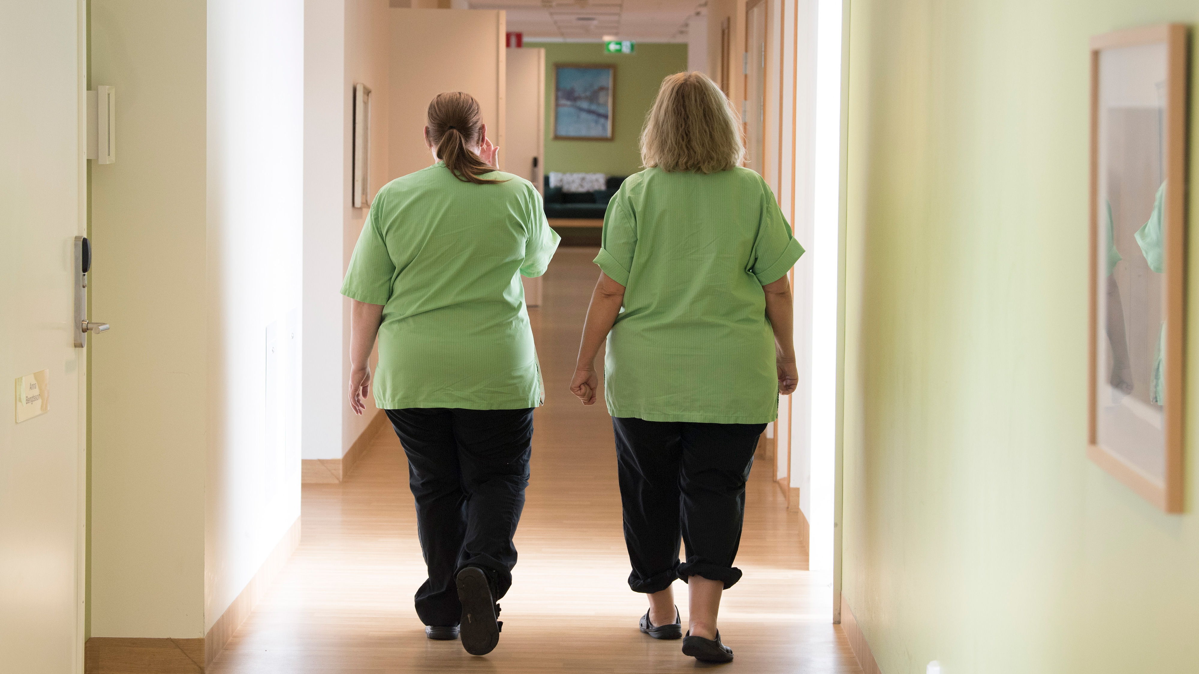Två kvinnor med likadana gröna skjortor och svarta byxor går i en korridor med ljusgröna väggar och trägolv, sedda bakifrån.