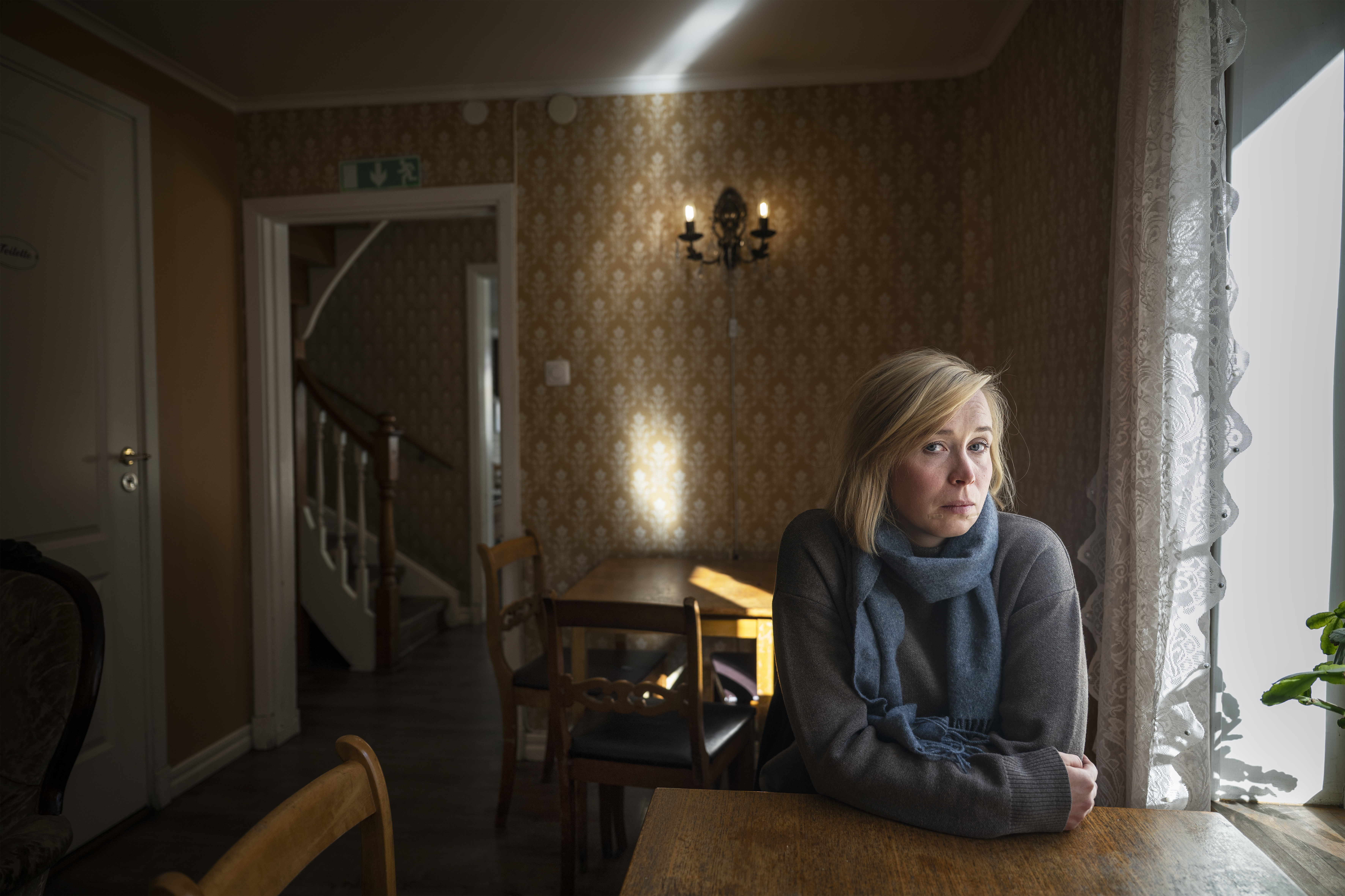 Kvinna med blont hår sitter vid ett bord vid ett fönster i ett rum med gula tapeter.