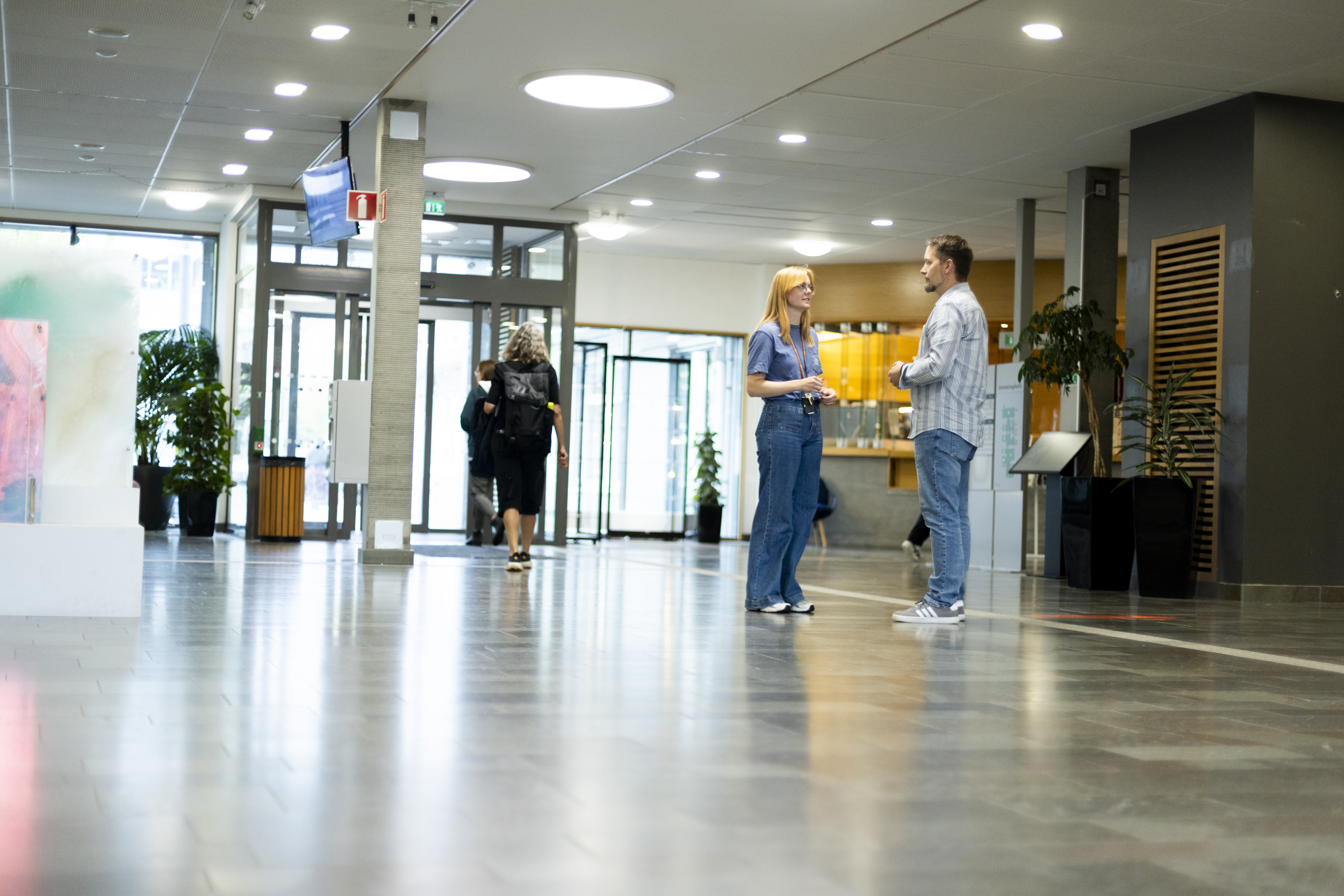 Stefan Lundgren står och pratar med en annan person i en byggnadsfoajé med glasdörrar, växter och andra som går i bakgrunden.