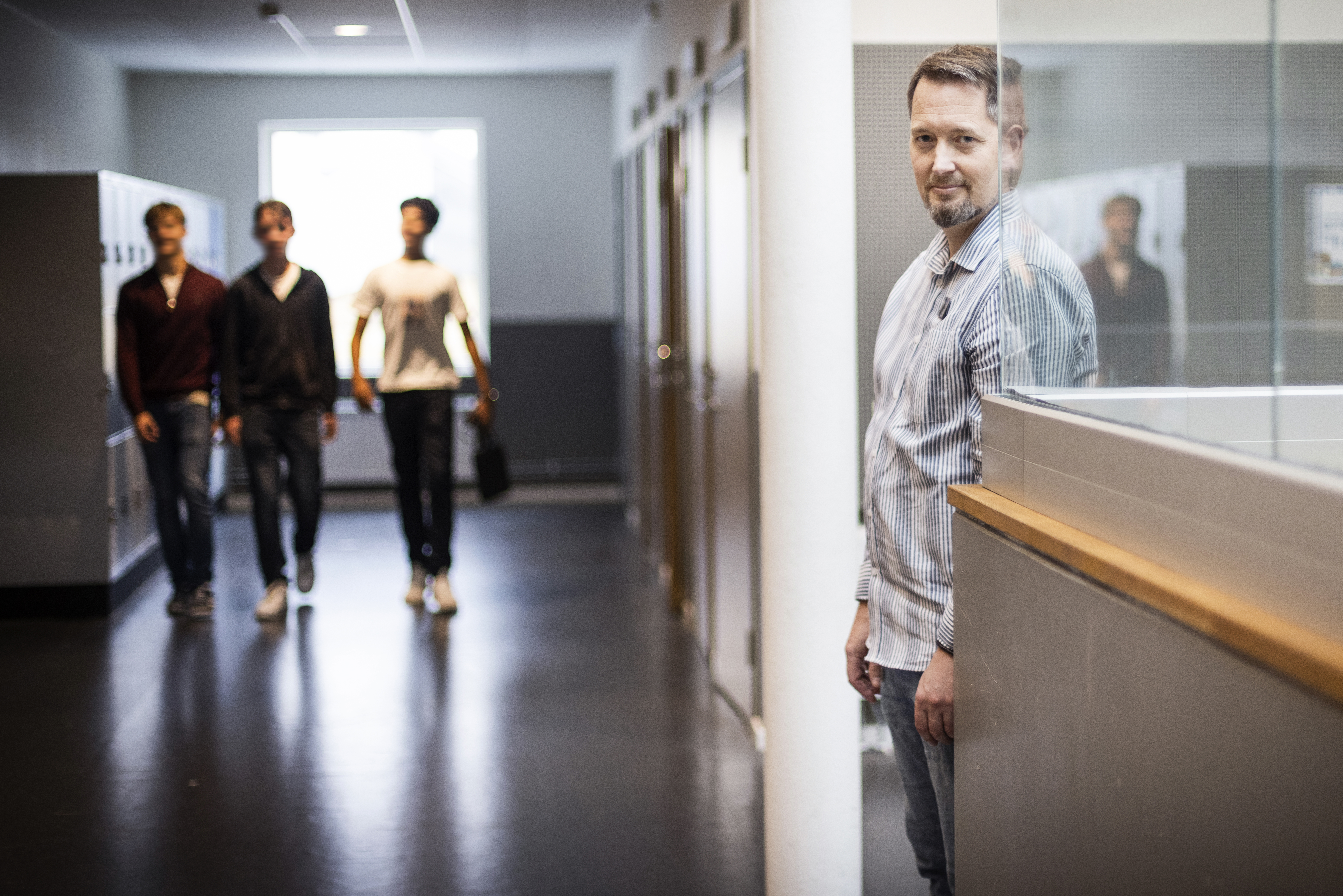 Stefan Lundgren står vid en glasvägg i en korridor, medan tre elever går tillsammans i bakgrunden.