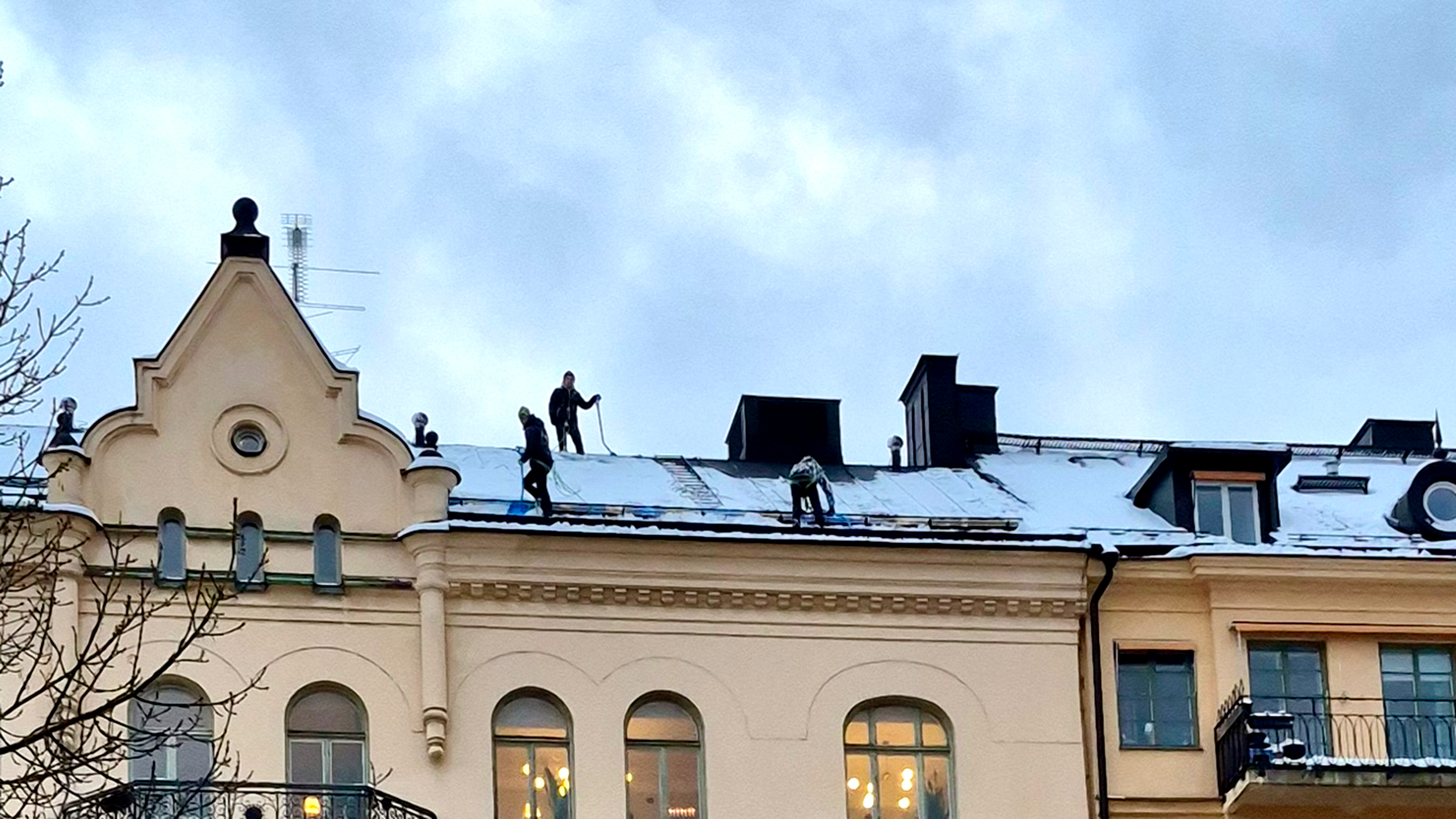 Tre arbetare skottar snö från taket på en beige byggnad under en molnig himmel.