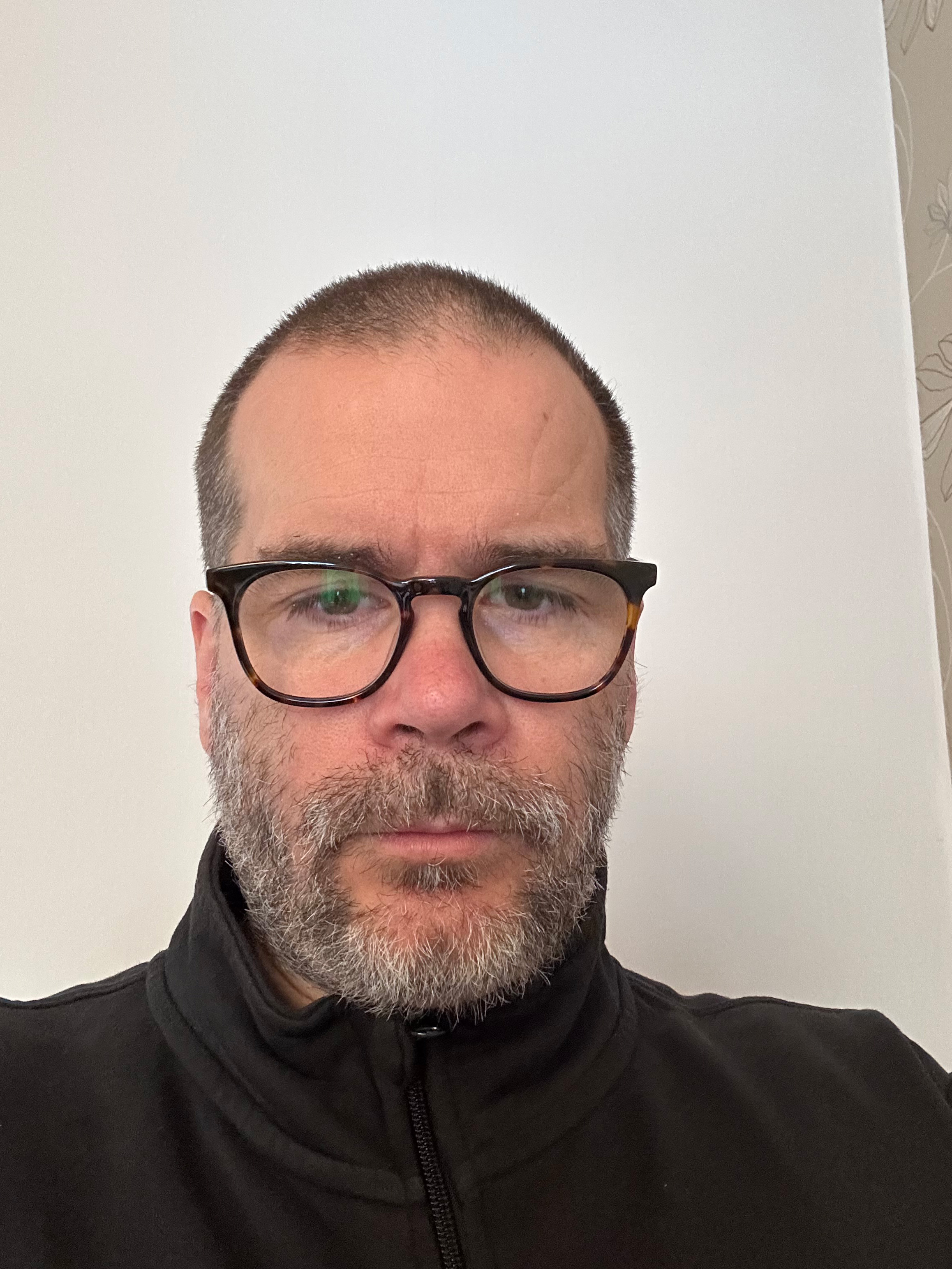 En man med kort hår, glasögon och skägg bär en svart jacka med dragkedja och står mot en vit vägg med mönster.