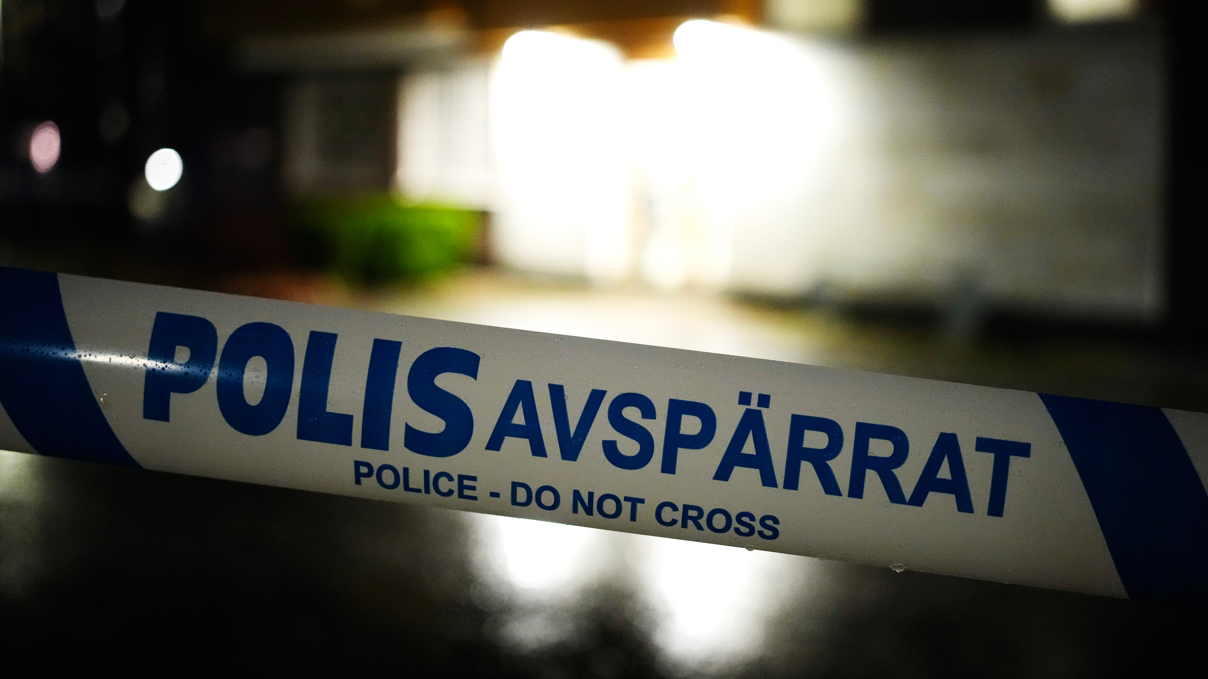 Närbild på svensk avspärrningstejp med texten "POLIS AVSPÄRRAT Police - Do Not Cross" framför en byggnad nattetid.