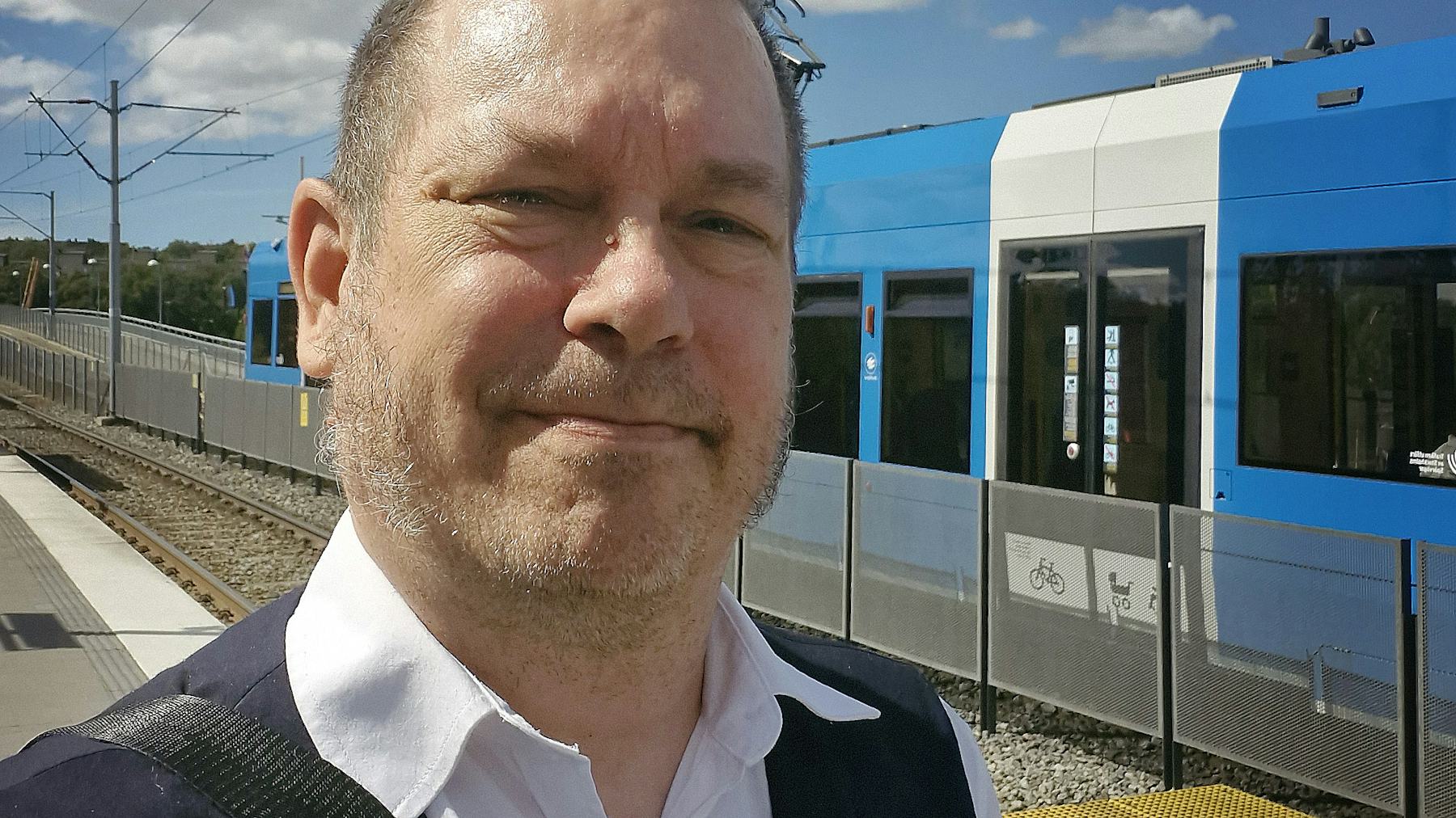 En man med skjorta står på en tågperrong. Ett blåvitt tåg syns i bakgrunden.
