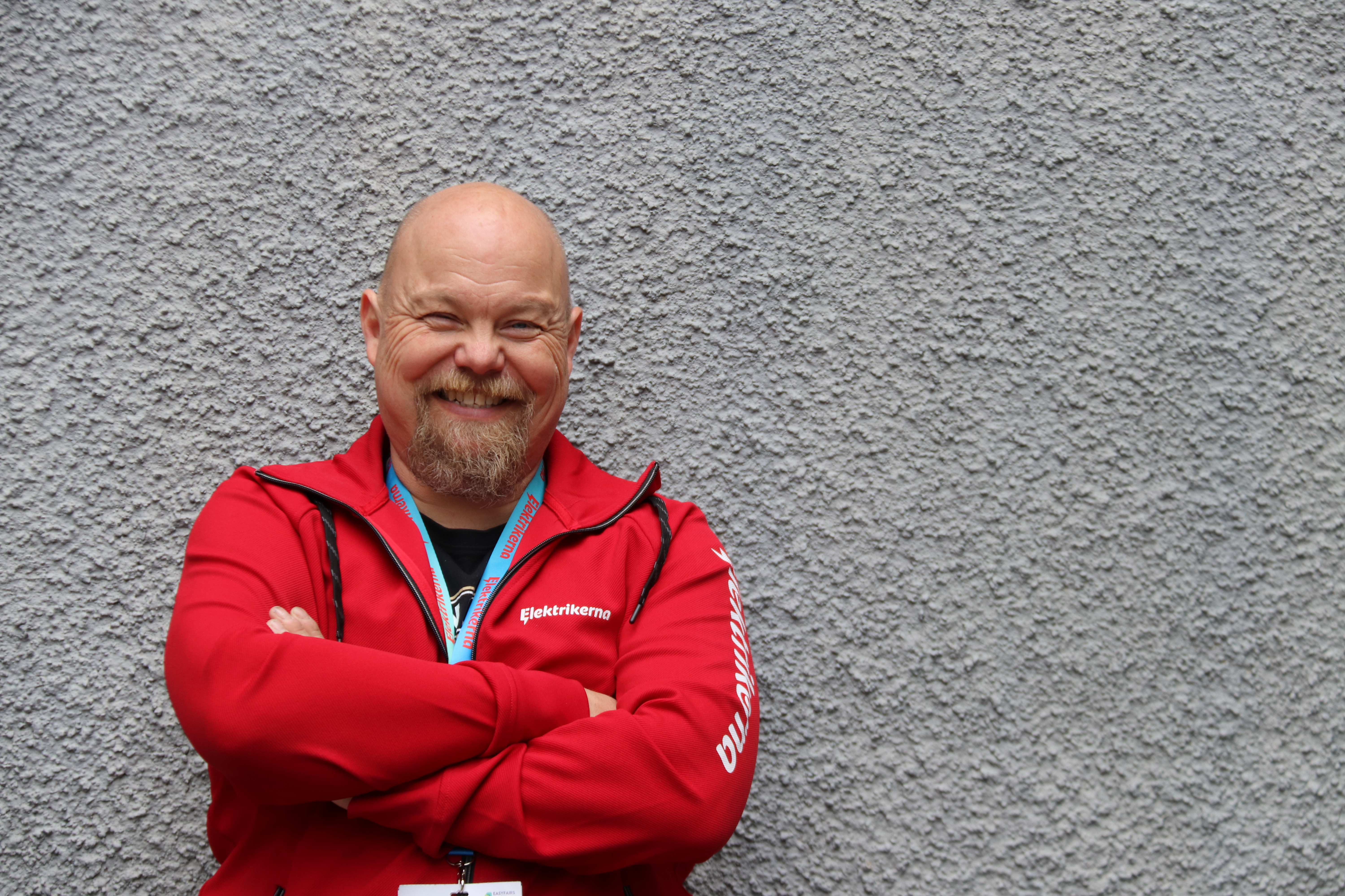 En skallig man med skägg står med armarna i kors, bär en röd jacka och ett nyckelband, framför en grå vägg.