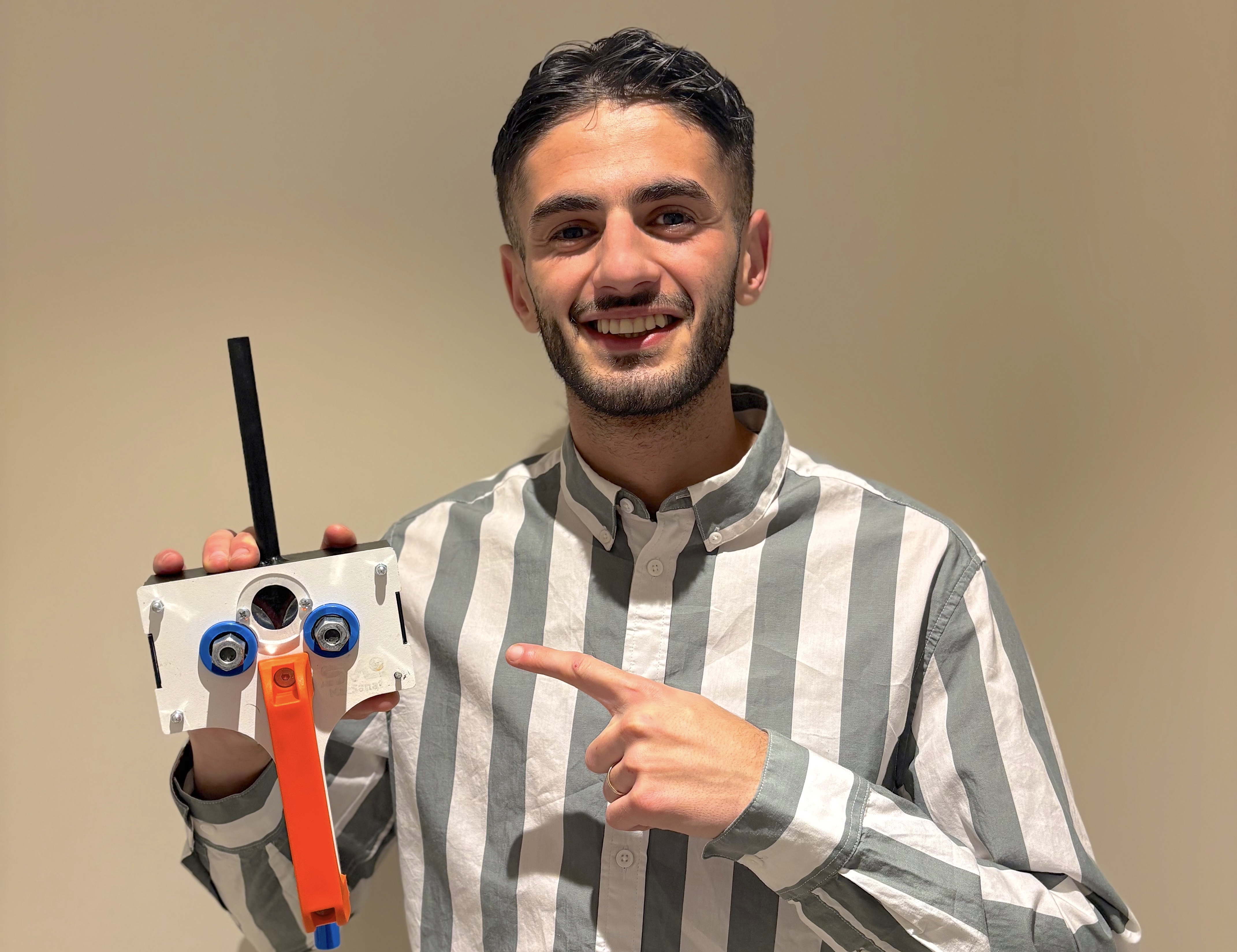 En man i randig skjorta pekar på ett handgjort robotliknande föremål med kugghjul och pinnar mot en vanlig bakgrund.