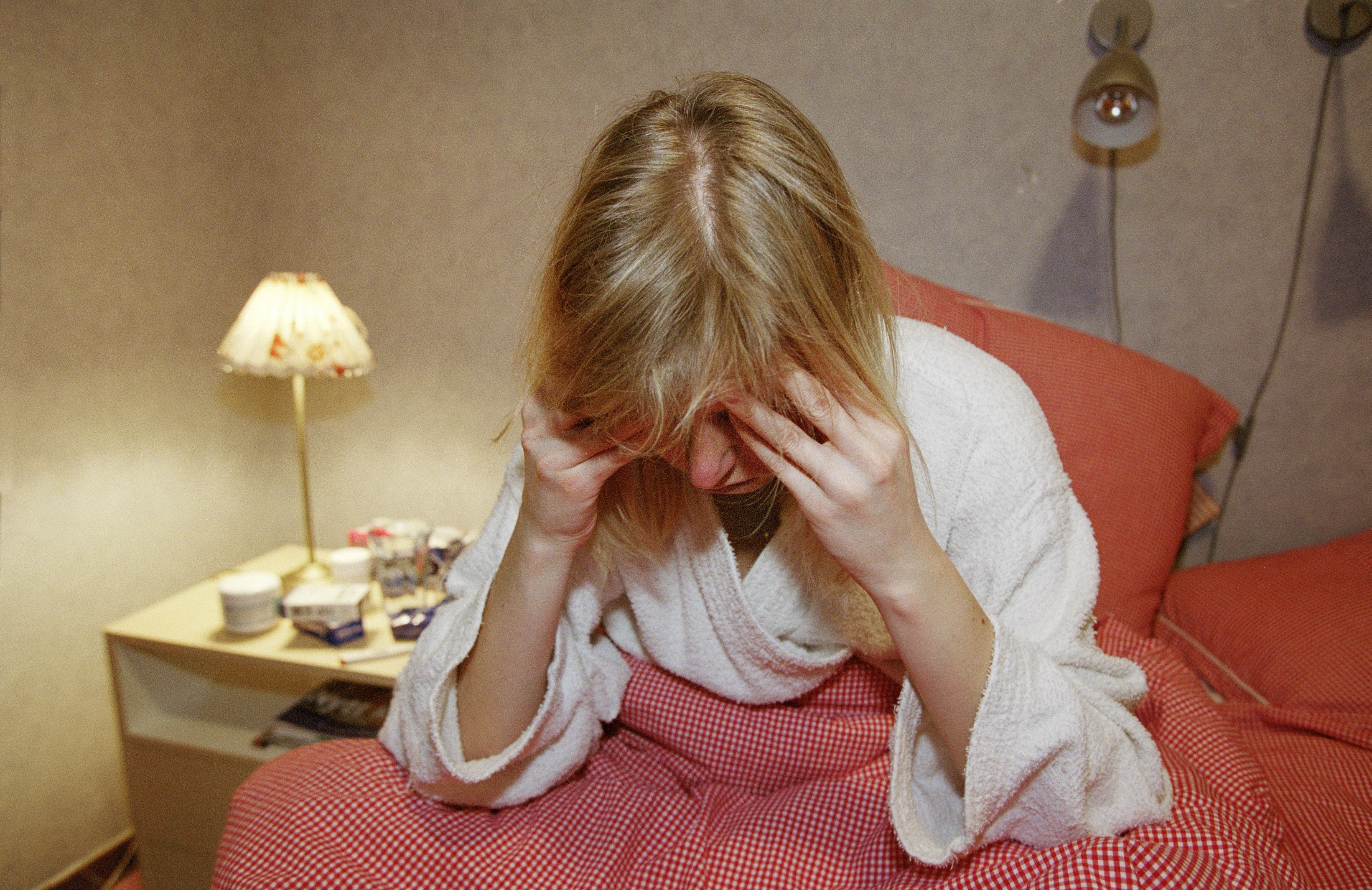 En person i vit morgonrock sitter på en säng med rött täcke och håller huvudet i händerna. En lampa står på nattduksbordet.
