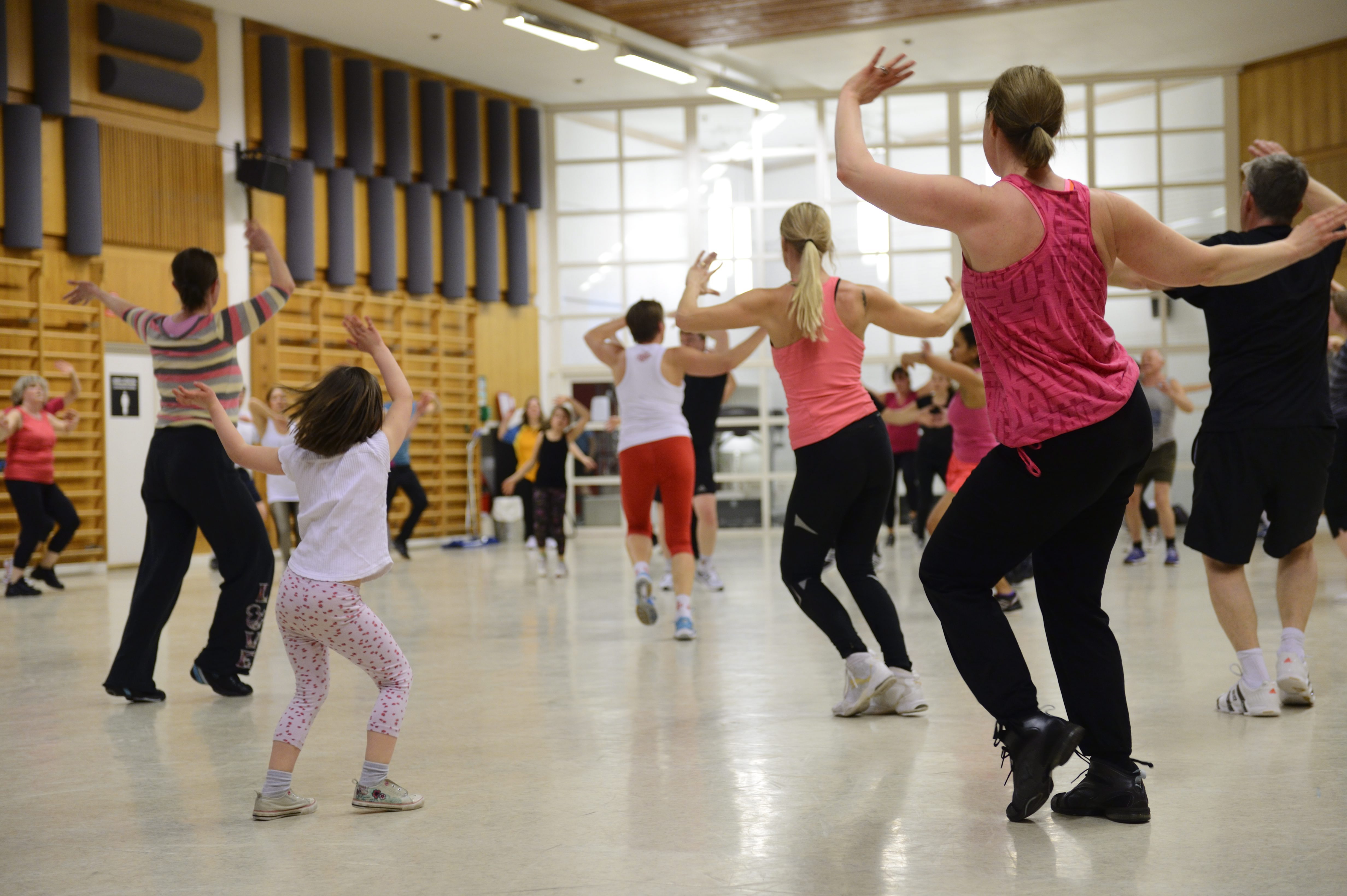 En grupp personer, både barn och vuxna, deltar i en tränings- eller dansklass inomhus och står vända framåt med armarna uppåt.