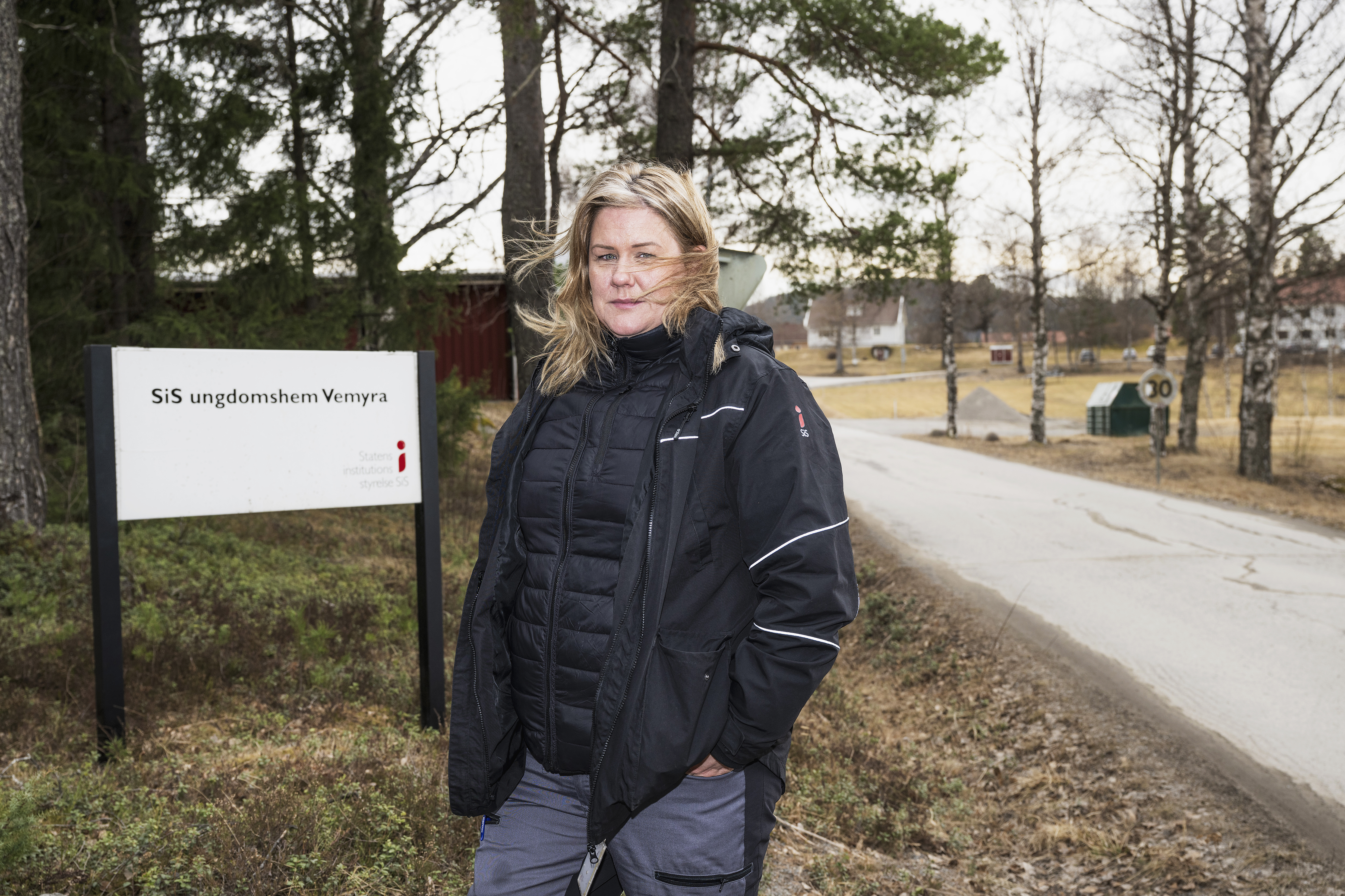 En kvinna i svart jacka står vid en väg bredvid en skylt som det står "SiS ungdomshem Vemyra" på, med träd och byggnader bakom.