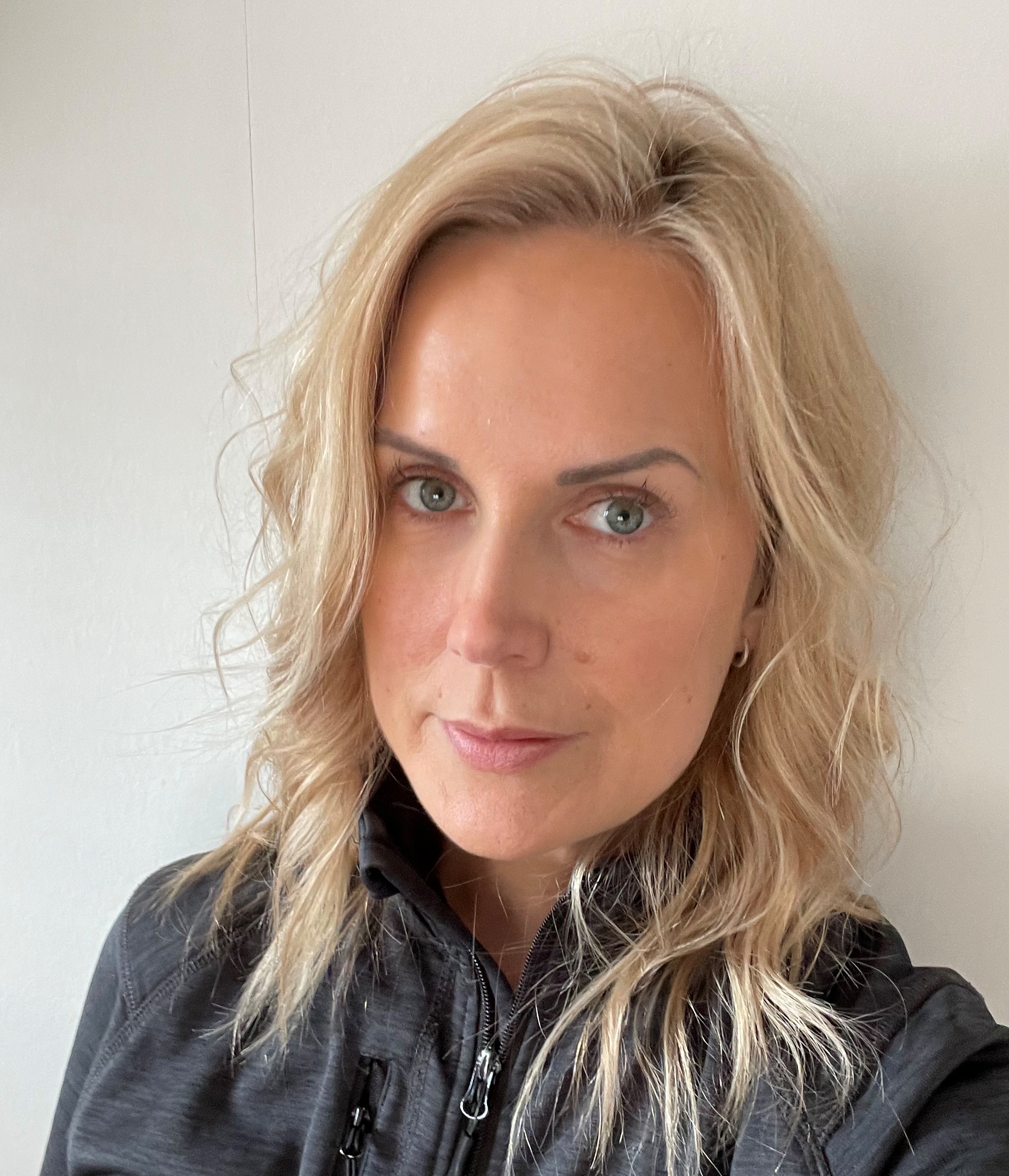 En kvinna med blont hår har på sig en mörk jacka med dragkedja och ser mot kameran mot en ljus bakgrund.