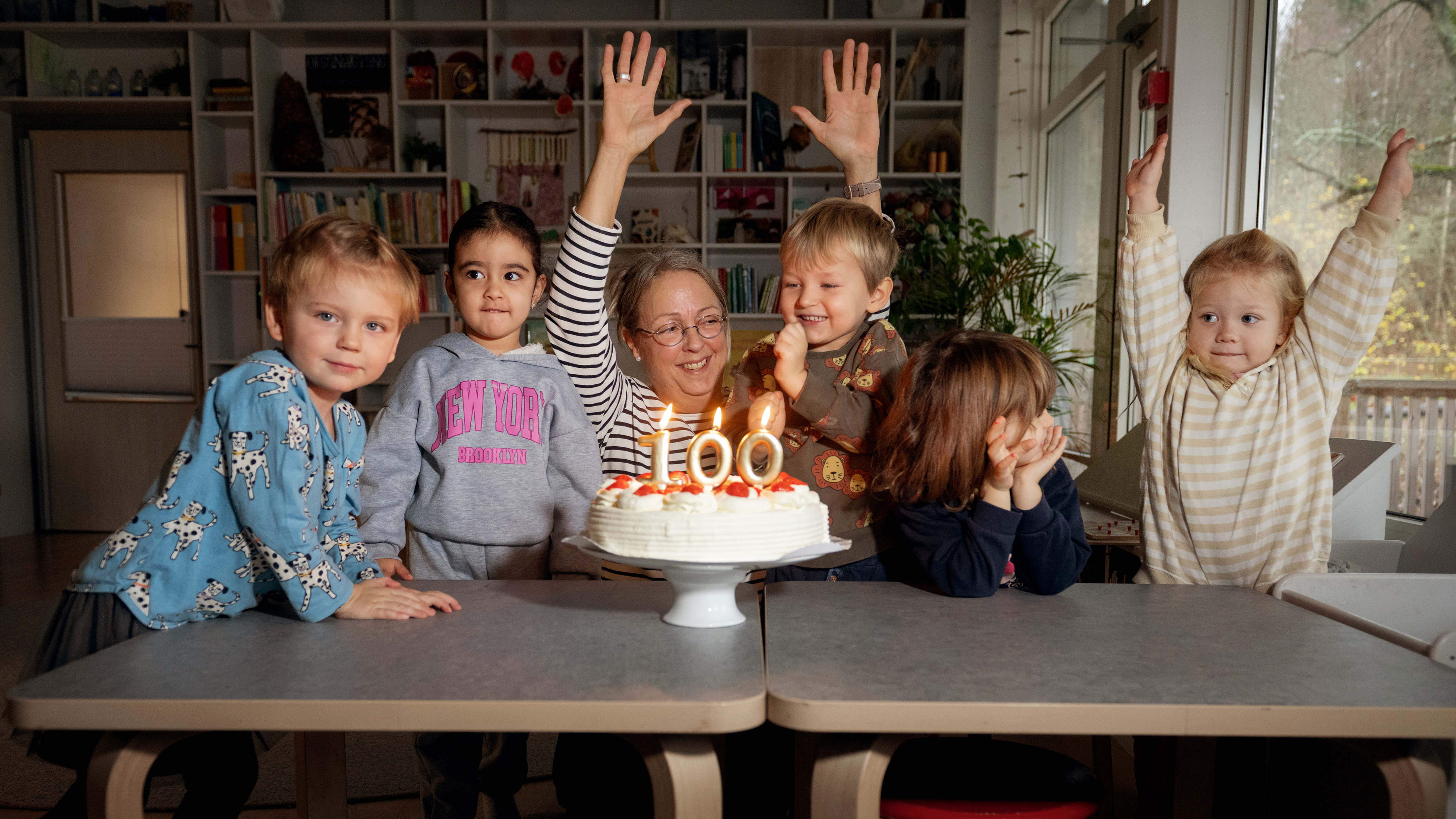 En vuxen och fem små barn sitter vid ett bord med en tårta med tända "100"-ljus; några barn räcker upp händerna och ler.