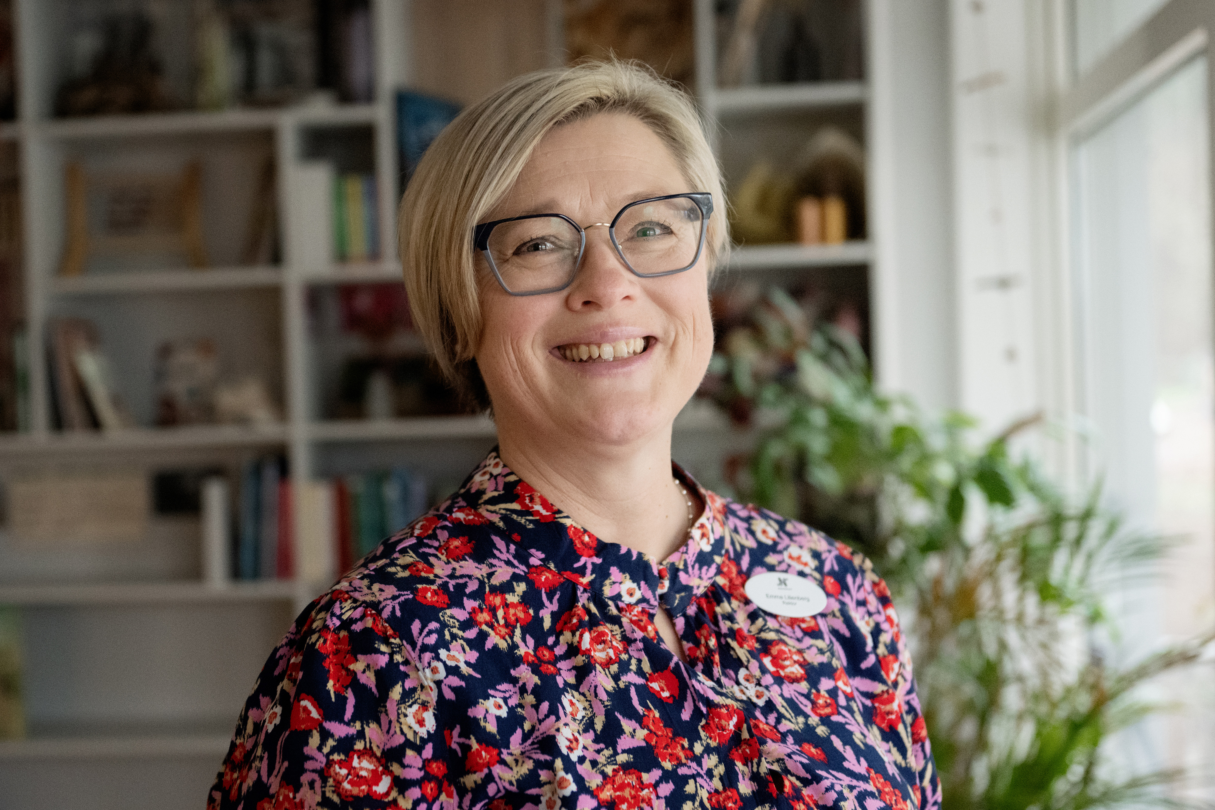 En kvinna med kort blont hår, glasögon och blus med blommönster står inomhus vid ett fönster med bokhyllor och växter.