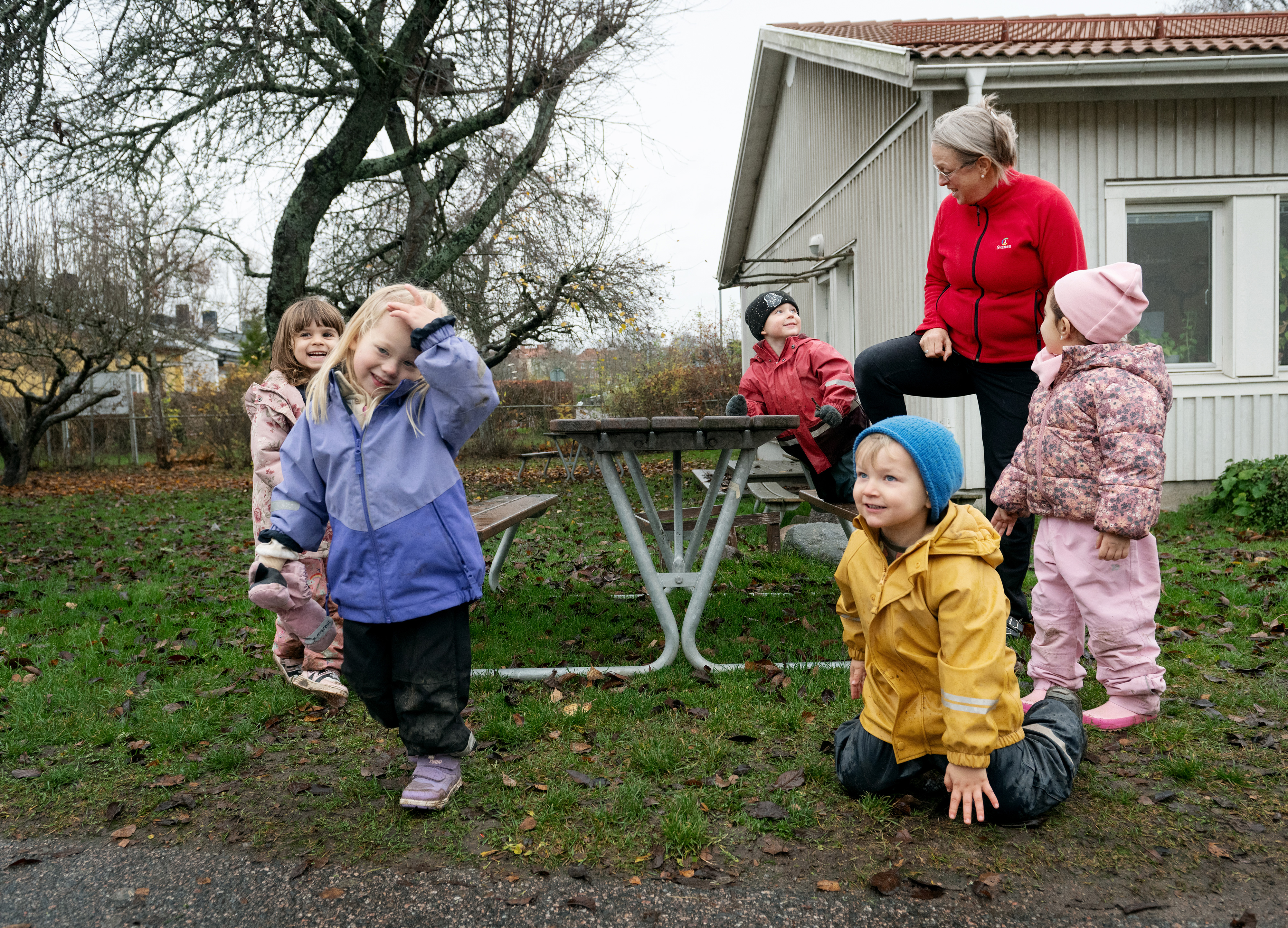 En grupp barn och en vuxen är utomhus vid ett träbord och en byggnad. Barnen har jackor på sig och leker på gräs och asfalt.