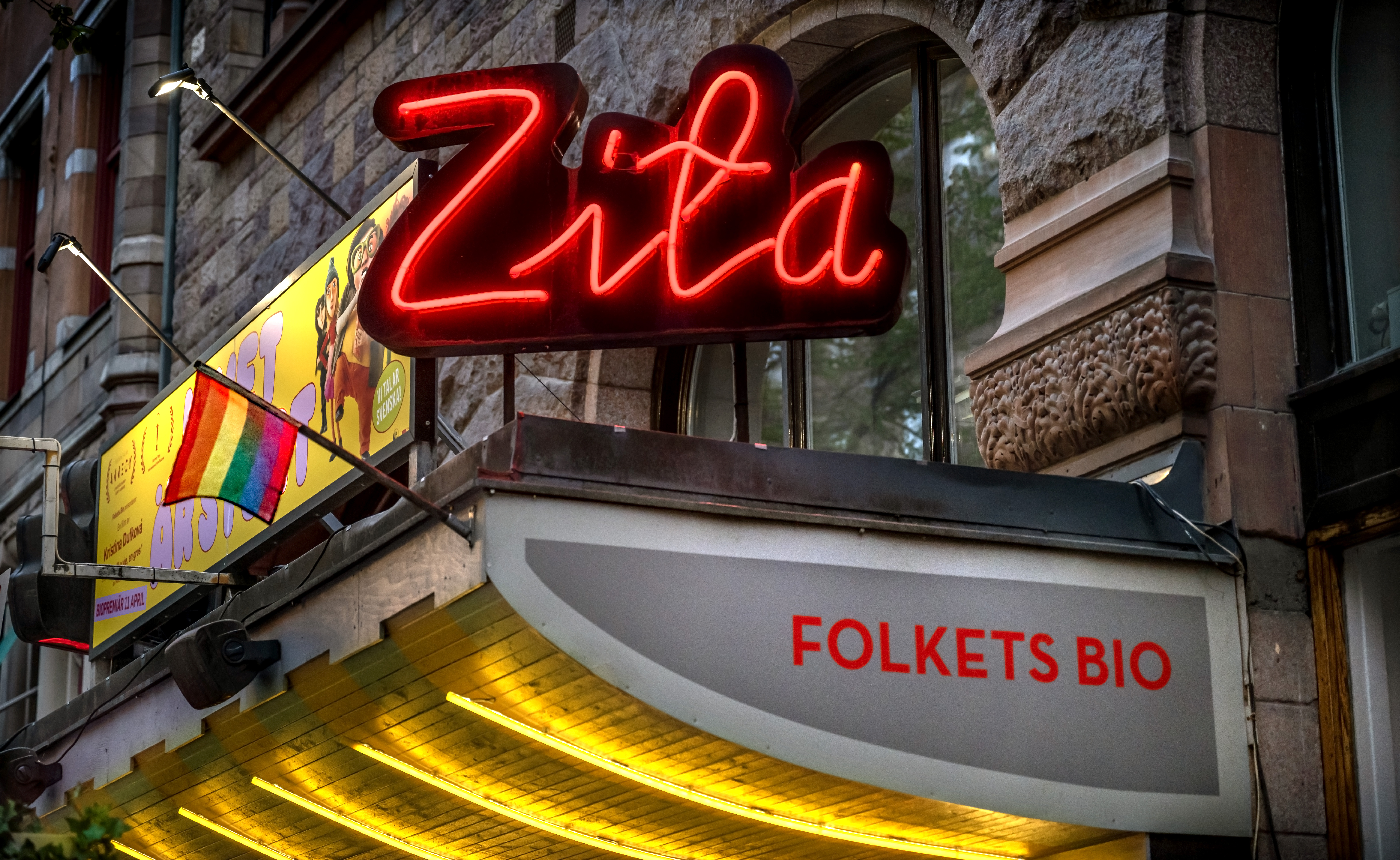 En neonskylt med texten "Zita" ovanför en biografsentré där "FOLKETS BIO" står i röda bokstäver, tegelvägg i bakgrunden.
