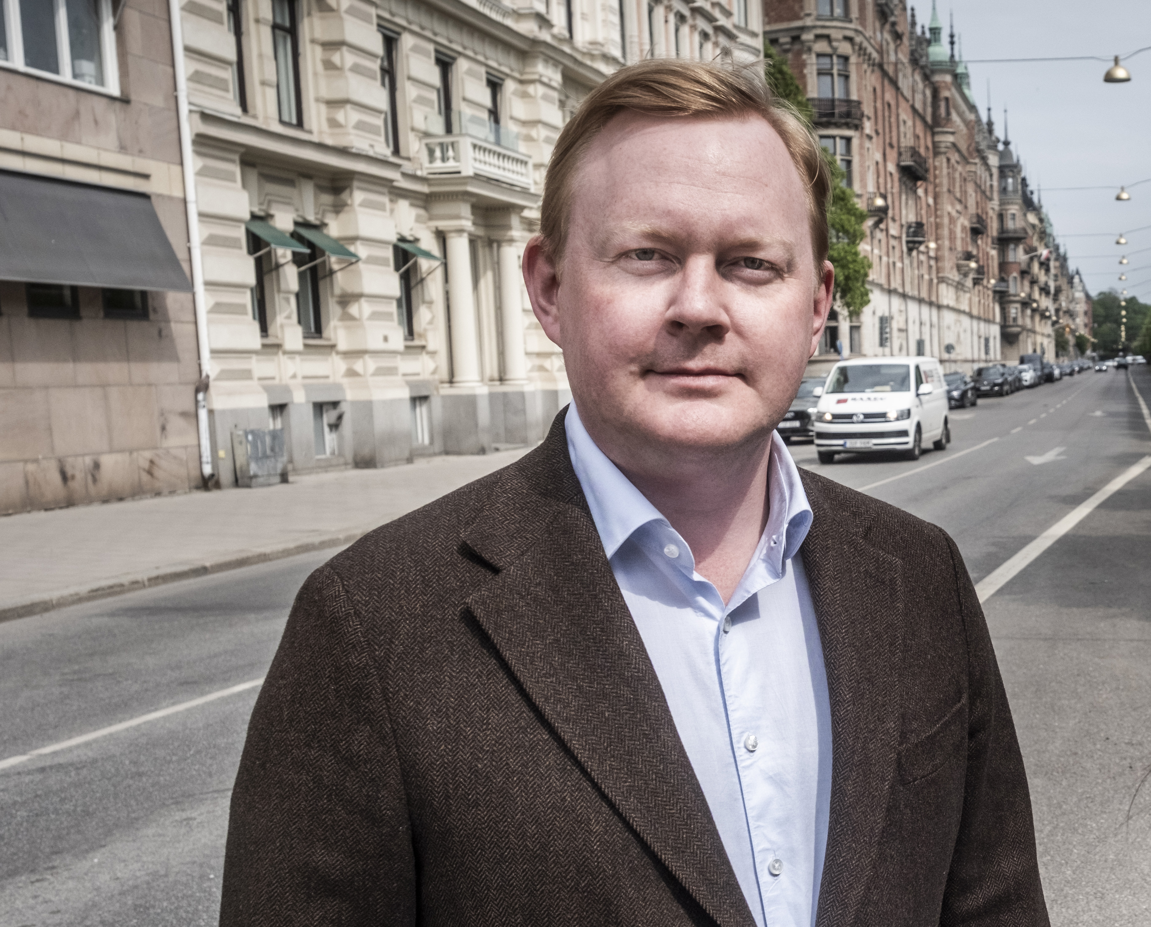 En man i brun kavaj och ljusblå skjorta står på en stadsgata med byggnader och parkerade bilar.