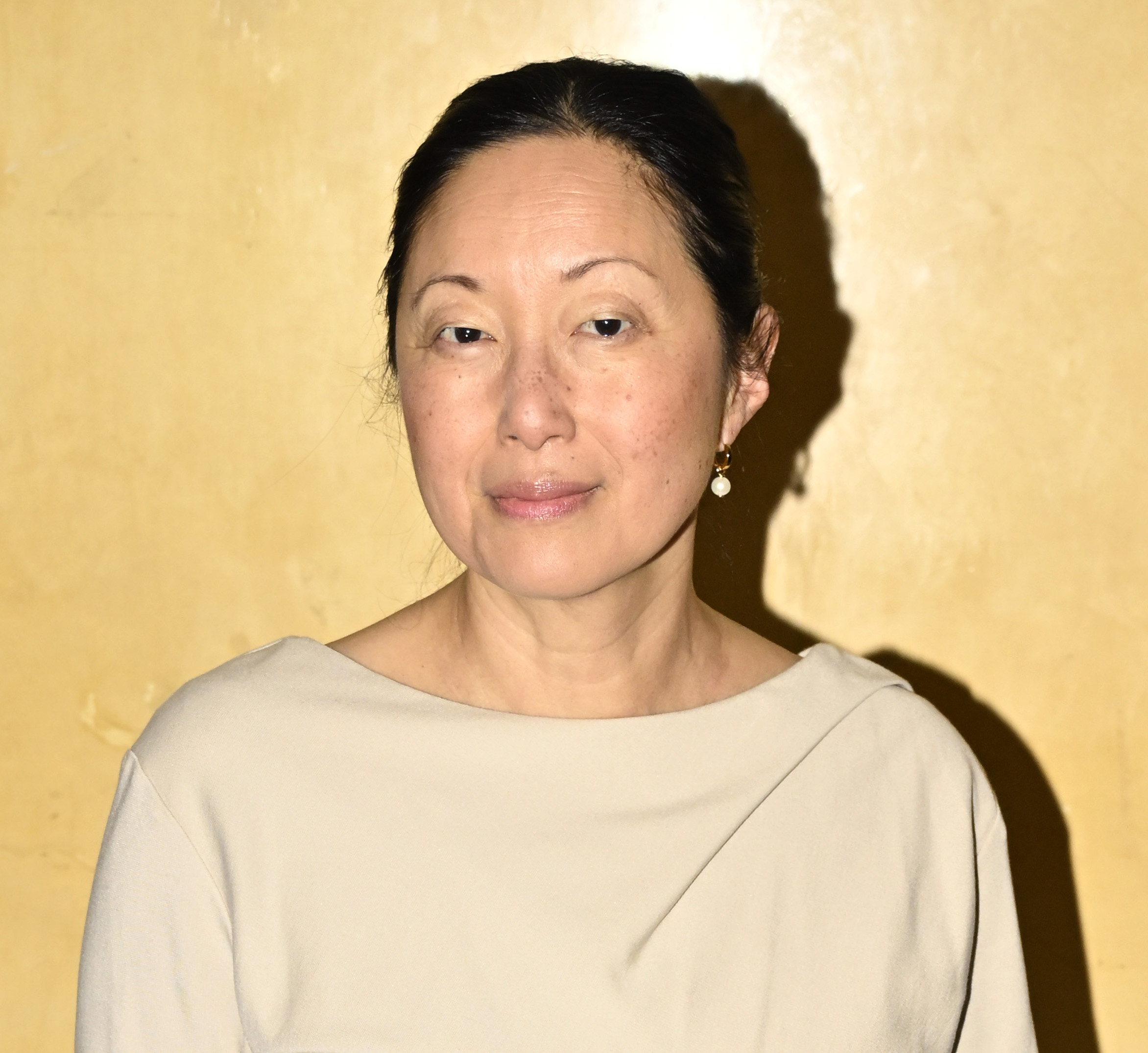 En kvinna med uppsatt mörkt hår har på sig en ljus topp och pärlörhängen, stående framför en gul vägg.