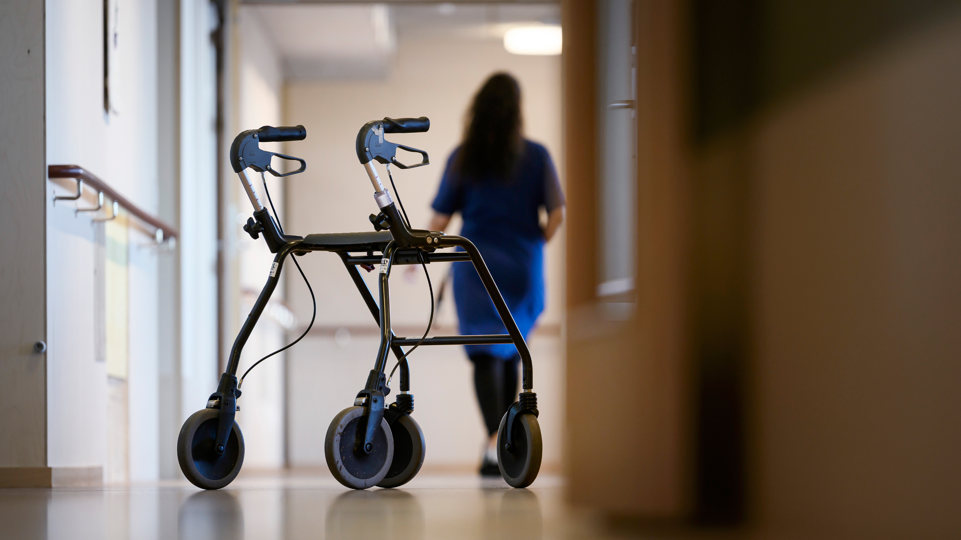 En rollator står i en korridor på en vårdanläggning, med en person i arbetskläder som går bortåt i bakgrunden.