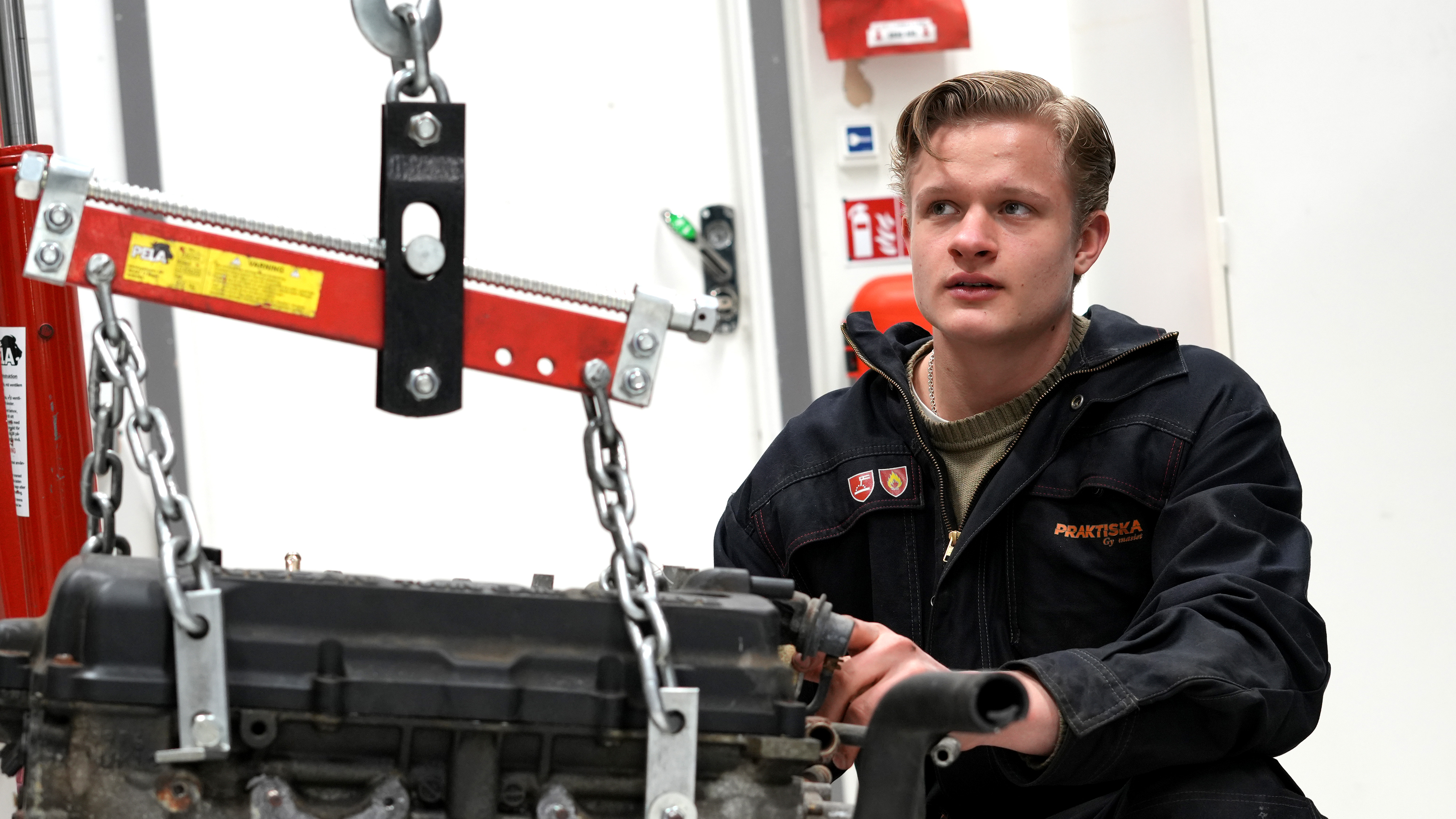 Ivar Andersson i mörk uniform arbetar med en motorlyft som är fäst vid en motor i en verkstad.