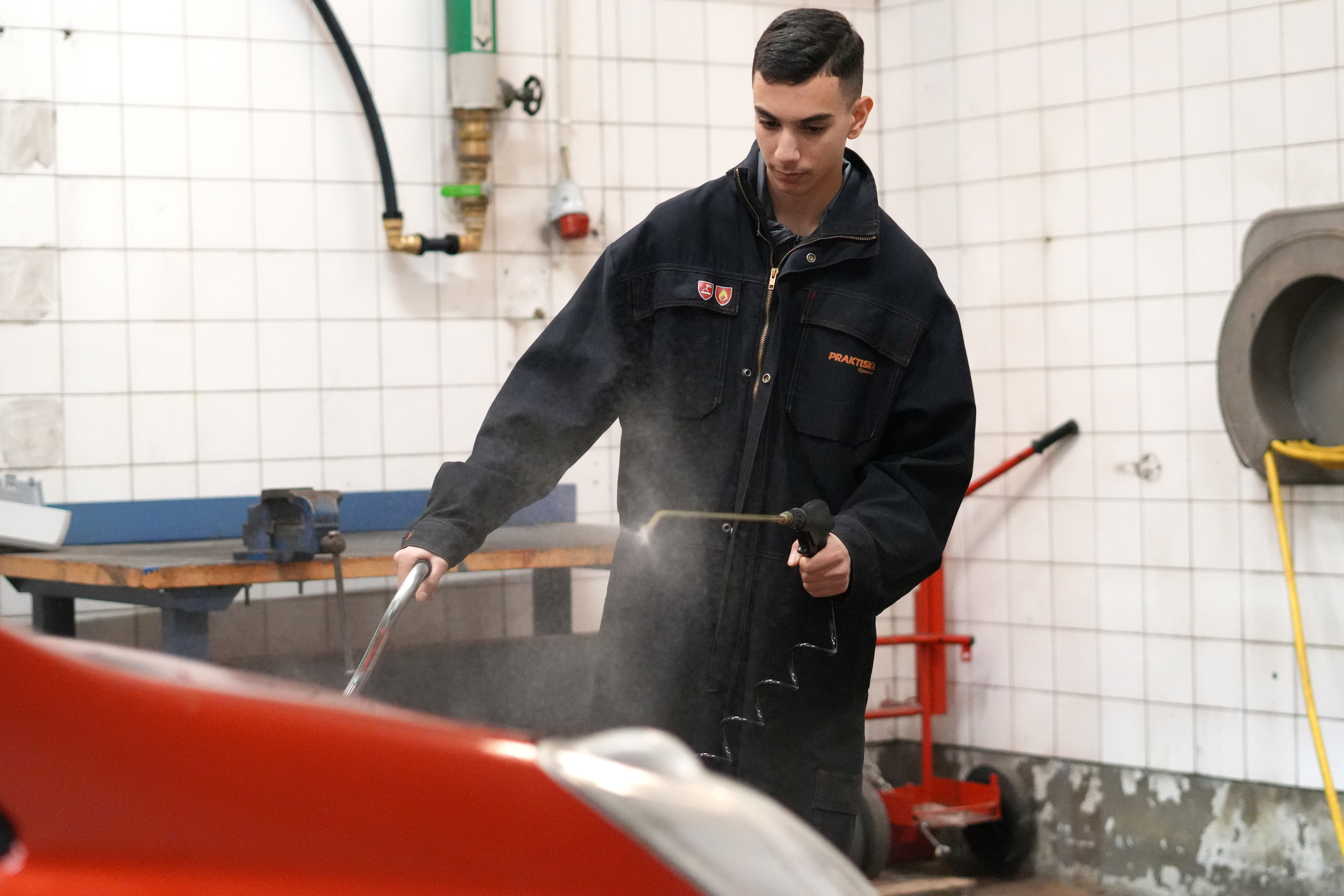 Hasan Hasanov i mörk jacka använder en högtryckstvätt för att tvätta ett rött fordon i en verkstad med kaklat golv.