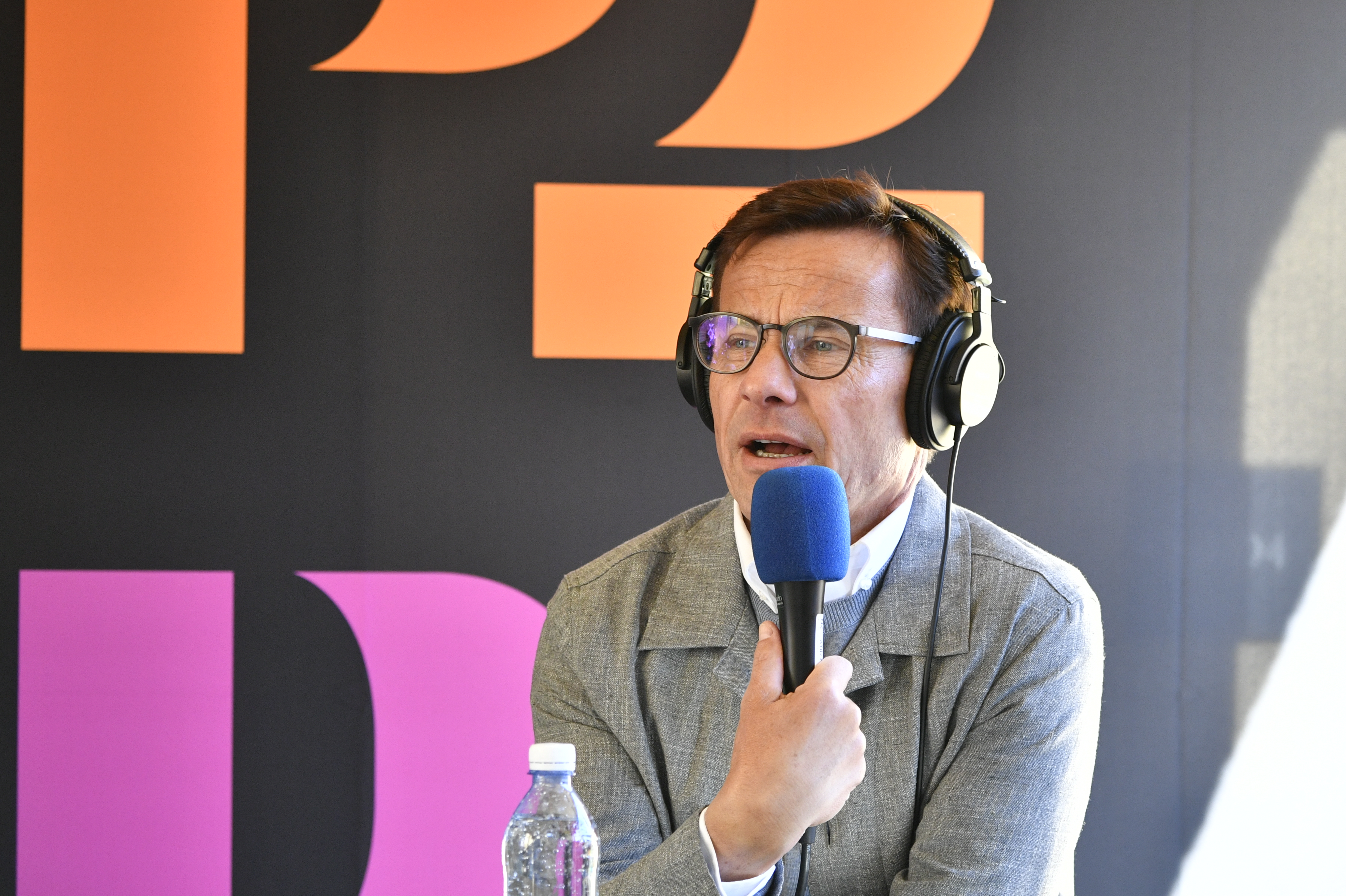 En man med hörlurar och glasögon pratar i en blå mikrofon. En vattenflaska står på bordet, bokstäver syns i bakgrunden.