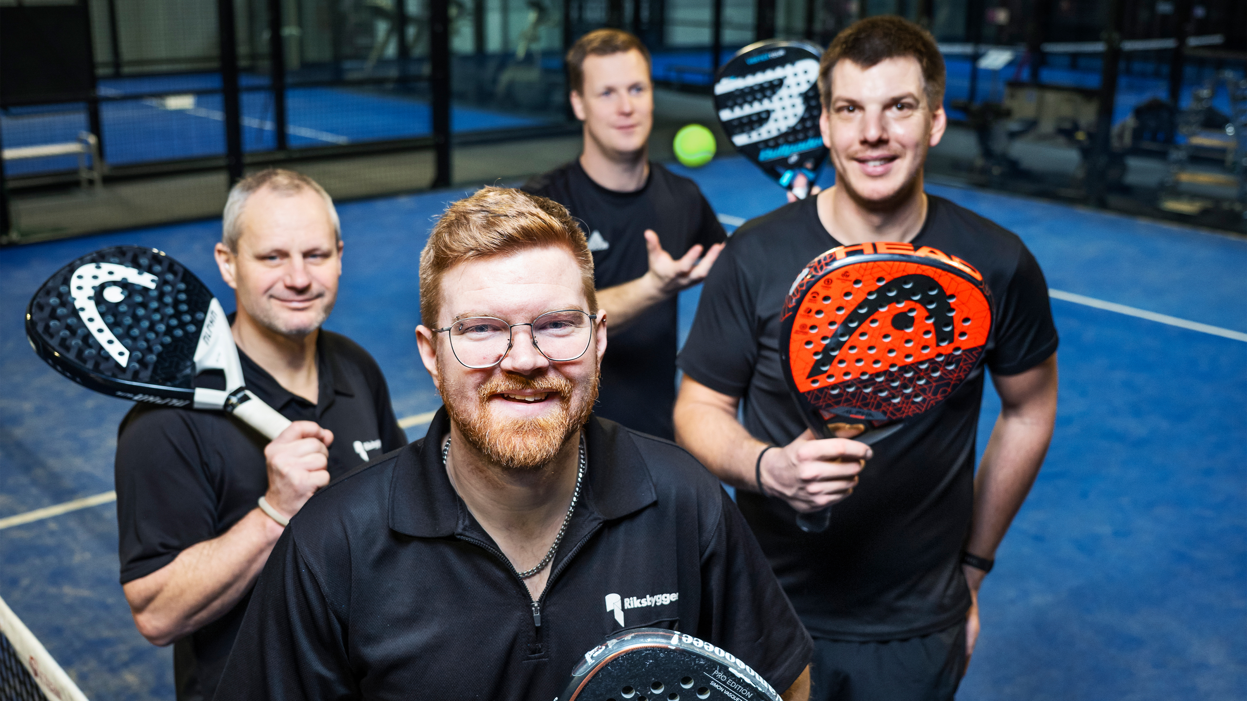 Fyra män i svarta tröjor håller padelracketar och står på en inomhusbana med blått underlag.