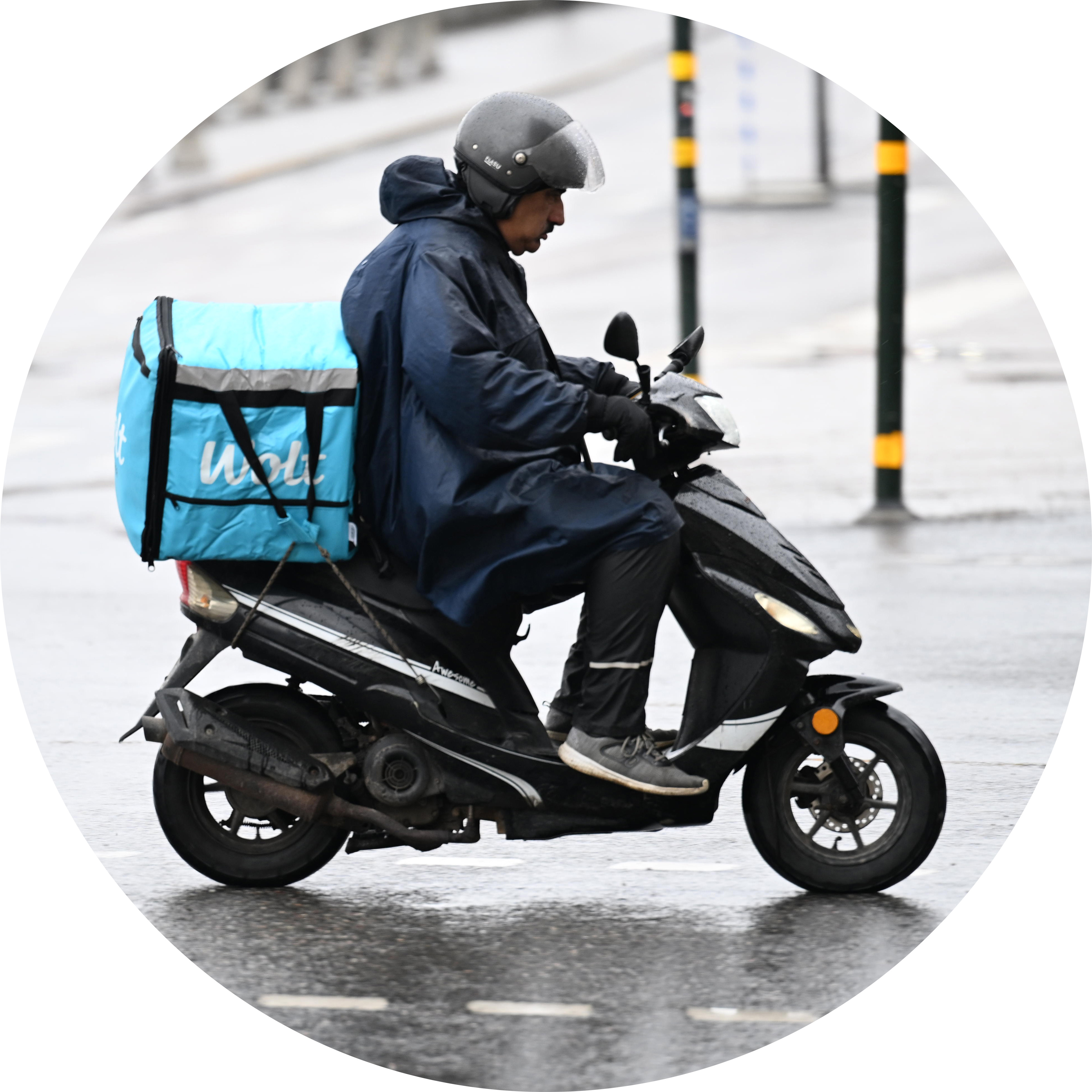 En matbud kör scooter med hjälm, regnjacka och en blå kylväska på en stadsgata som är blöt av regn.