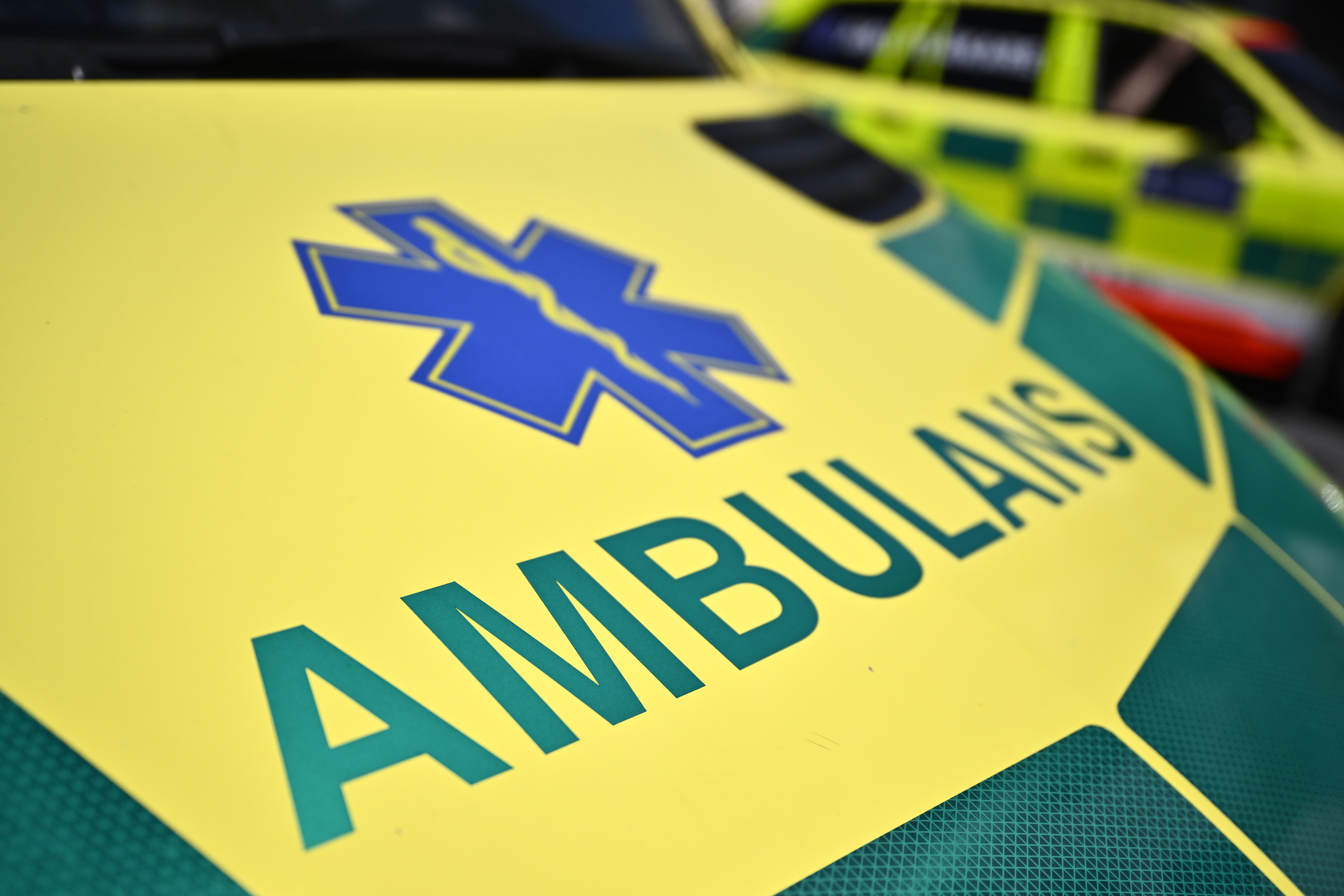 Närbild på ambulansens motorhuv med texten "AMBULANS" och en blå sjukvårdssymbol; ett annat utryckningsfordon i bakgrunden.