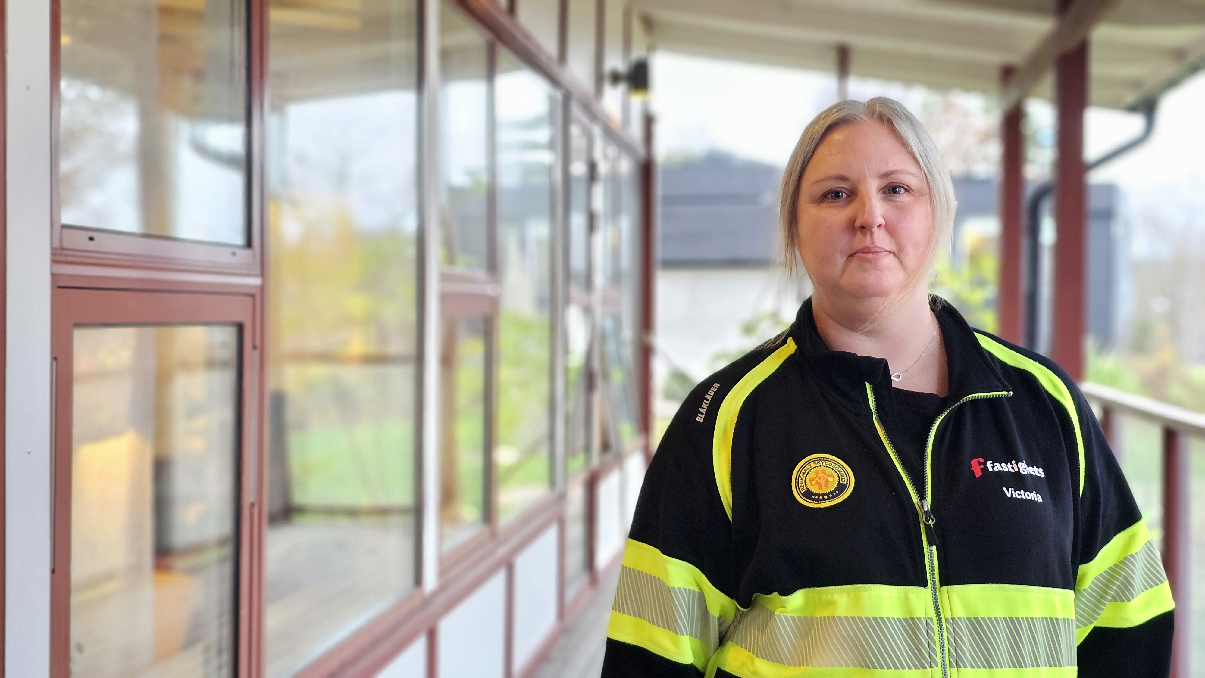 En kvinna klädd i en gul och svart tröja märkt med "Skyddsmedel" Stå utomhus nära en byggnad med stora fönster.