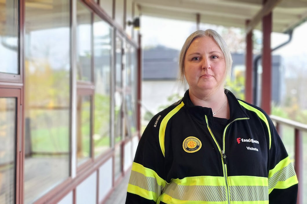 En kvinna klädd i en gul och svart tröja märkt med "Skyddsmedel" Stå utomhus nära en byggnad med stora fönster.