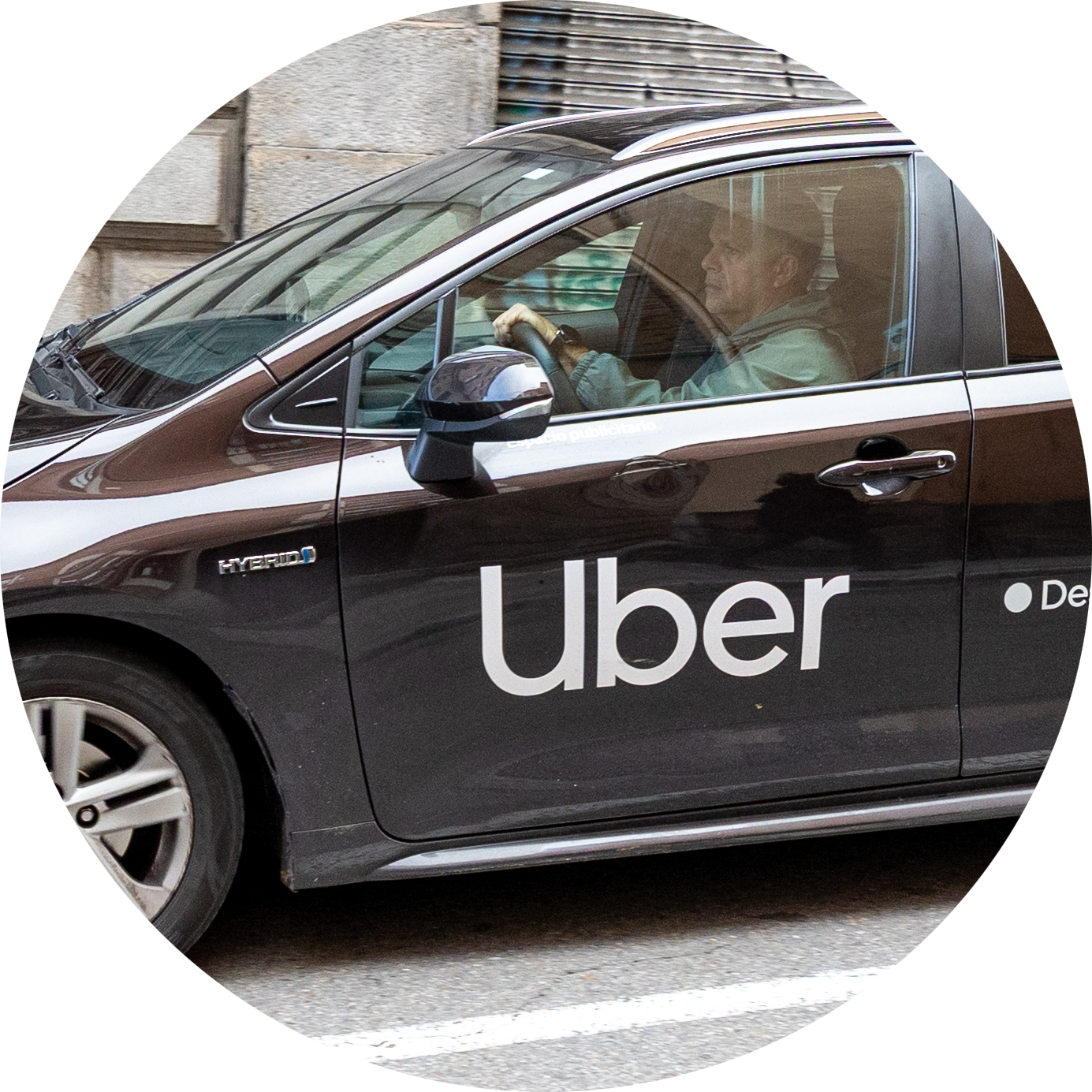 En man kör en svart Uber-märkt hybridbil på en stadsgata, sedd från sidan genom förarens fönster.