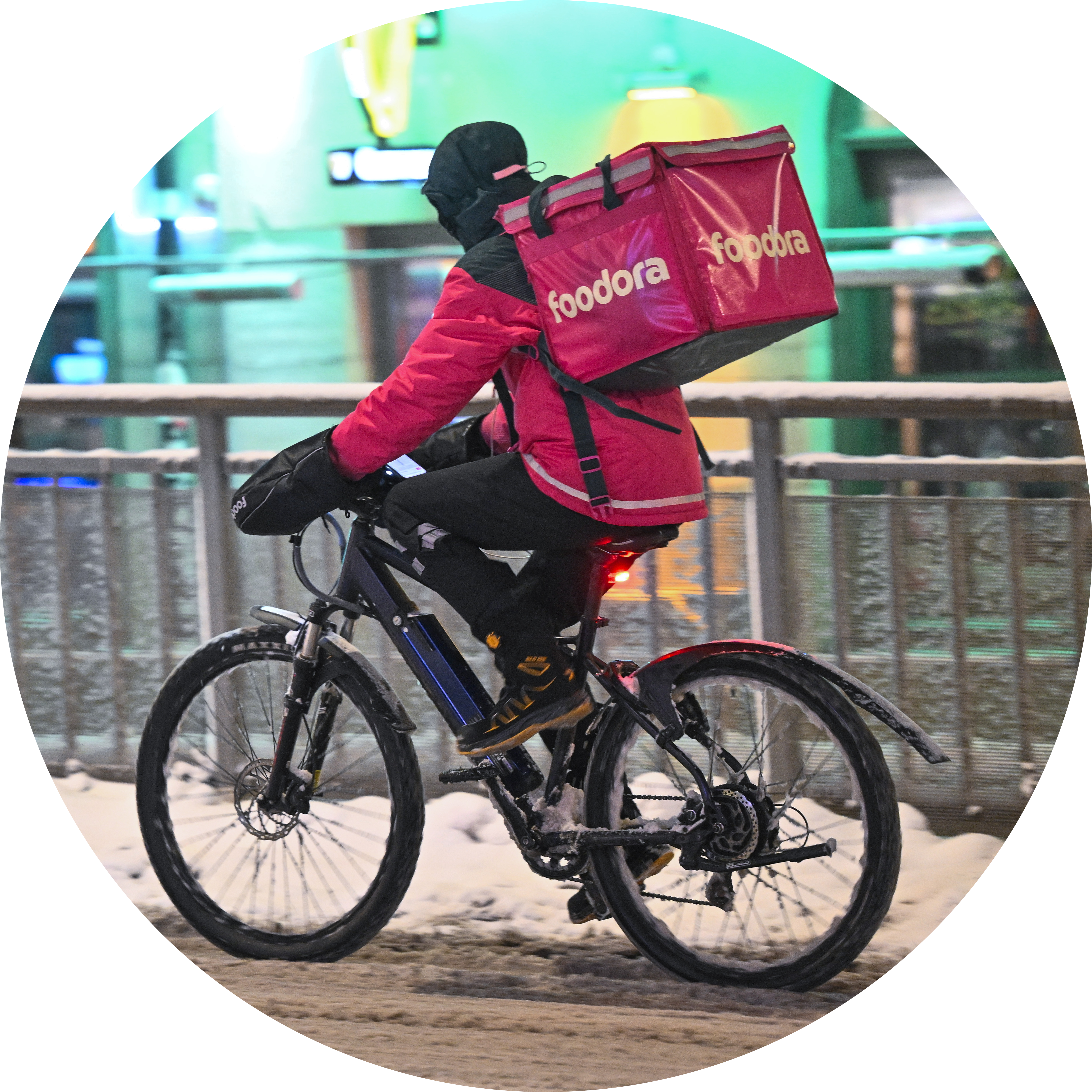 En person med vinterkläder cyklar på en snötäckt gata och bär en stor rosa Foodora-leveransväska.