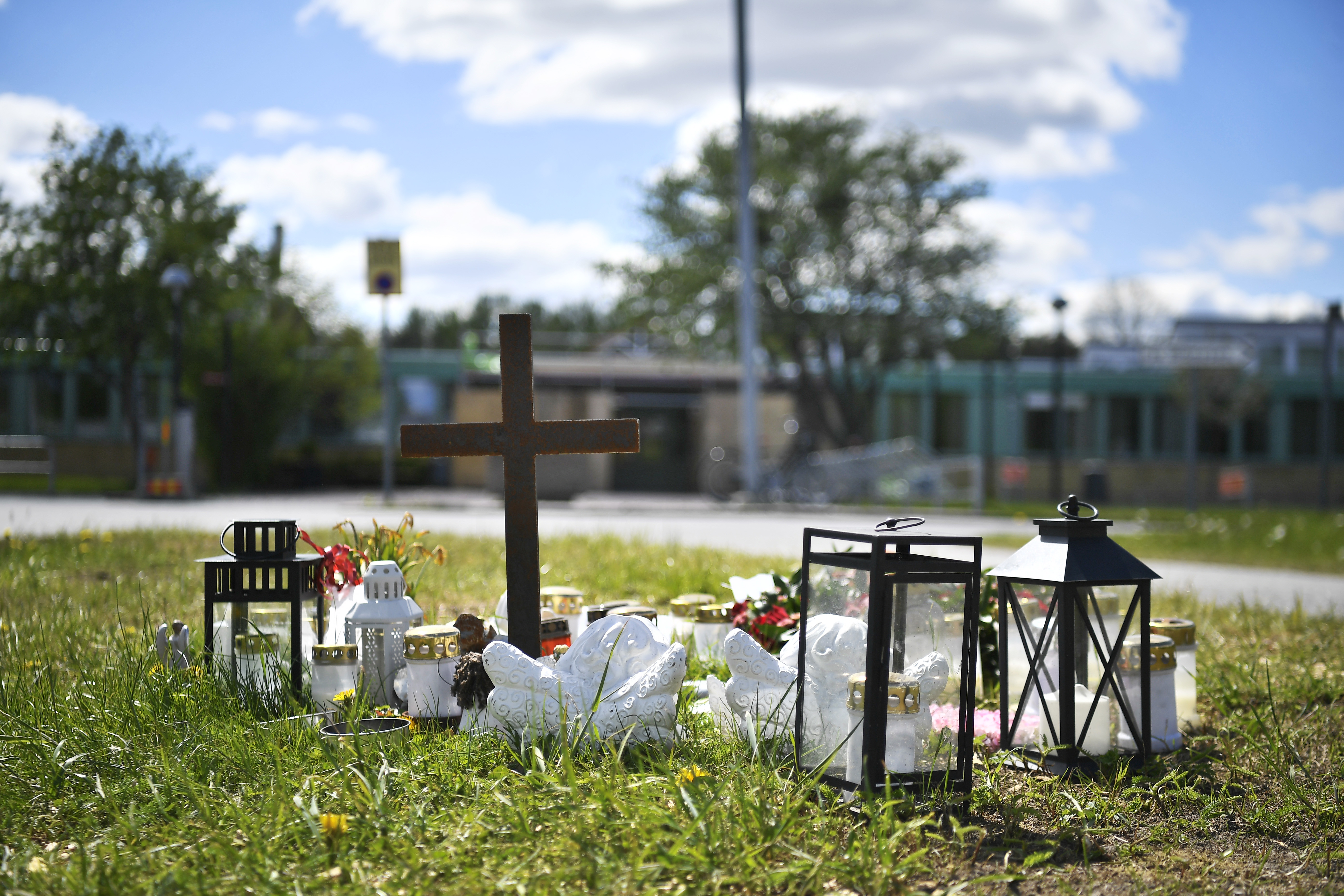 Ett träkors samt ljus, lyktor och blommor är placerade som ett minnesmärke på gräs utomhus med byggnader i bakgrunden.