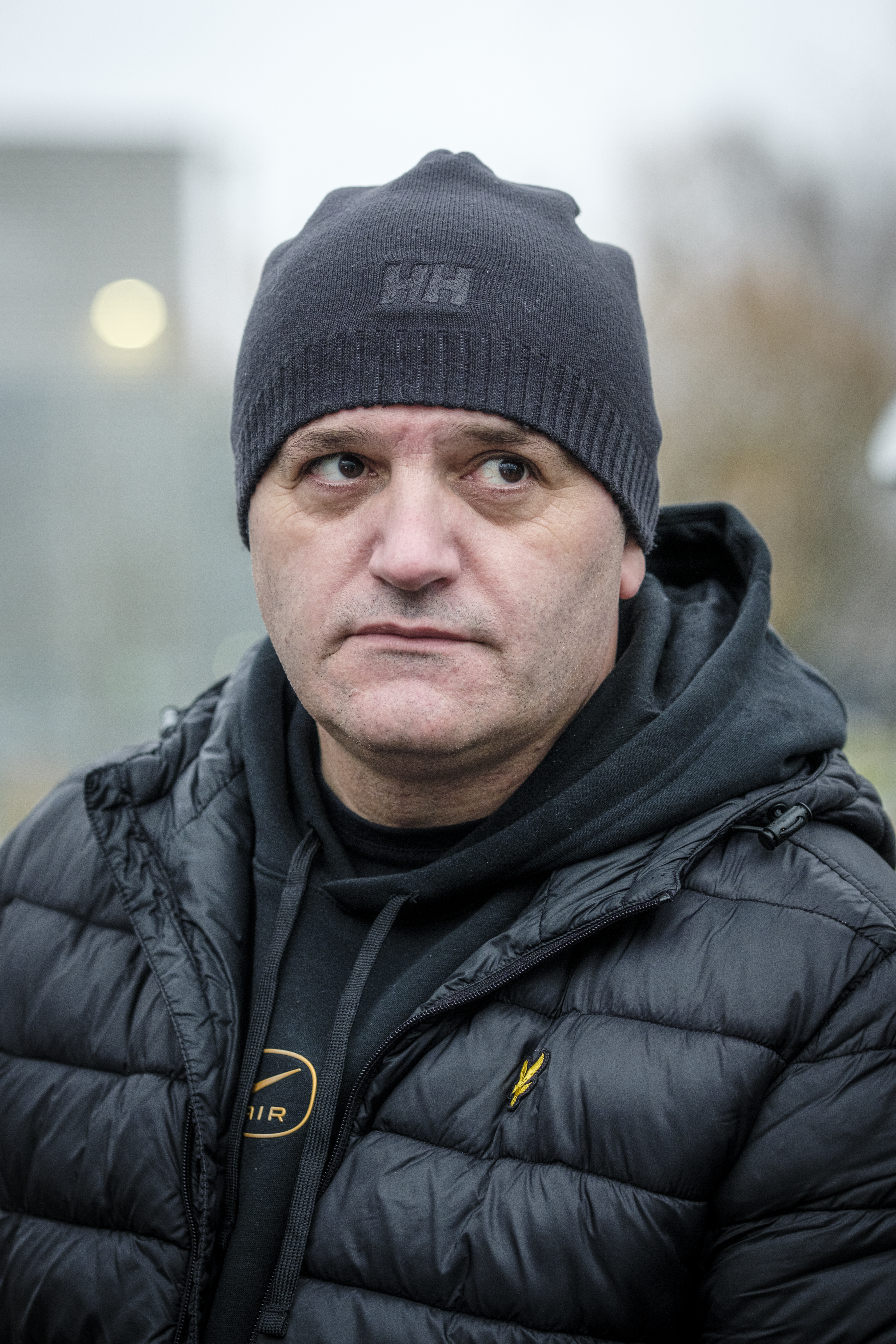 Muljhan med svart mössa, svart dunjacka och svart huvtröja står utomhus och tittar lite åt sidan med neutral min.