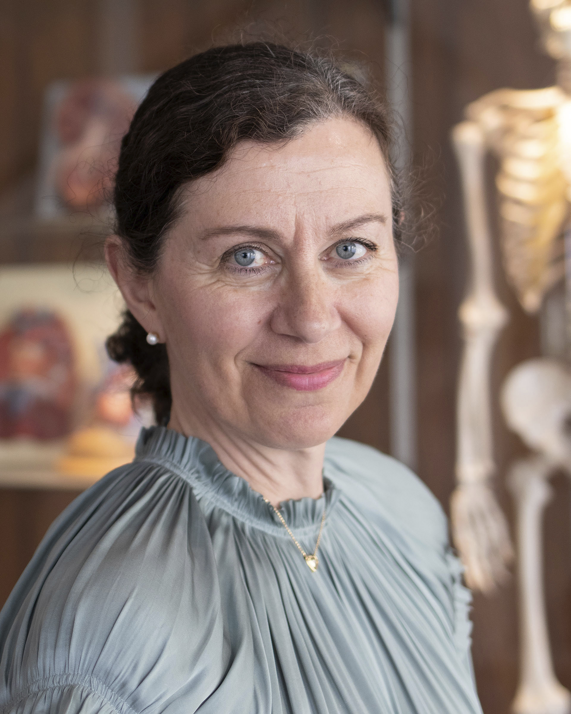 En kvinna med blå ögon och mörkt hår uppsatt, i en ljusgrå plisserad blus, står inomhus med en suddig anatomisk modell bakom sig.