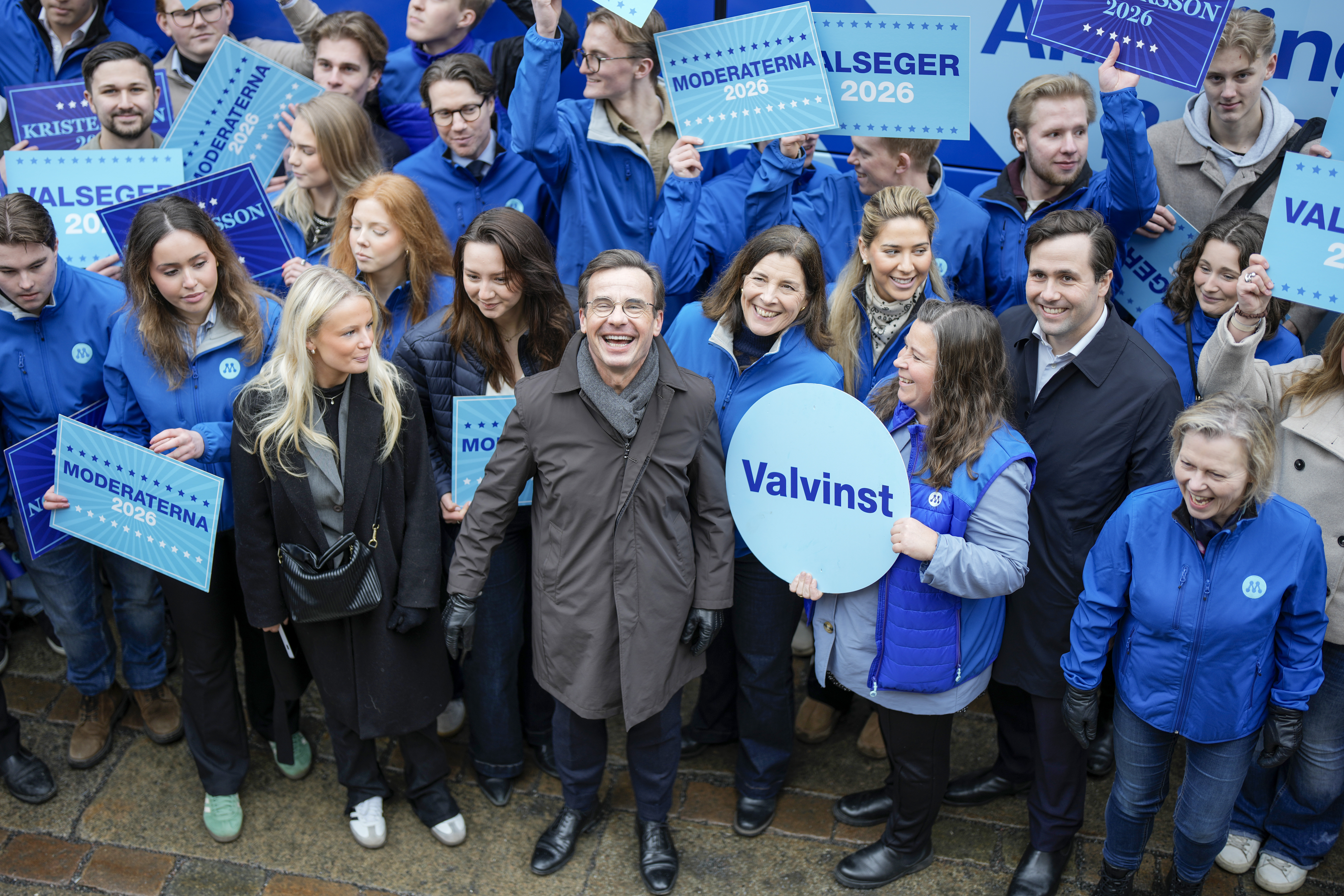 En grupp personer, flera med blå jackor, står tillsammans och håller upp skyltar med politiska budskap under ett kampanjevenemang.