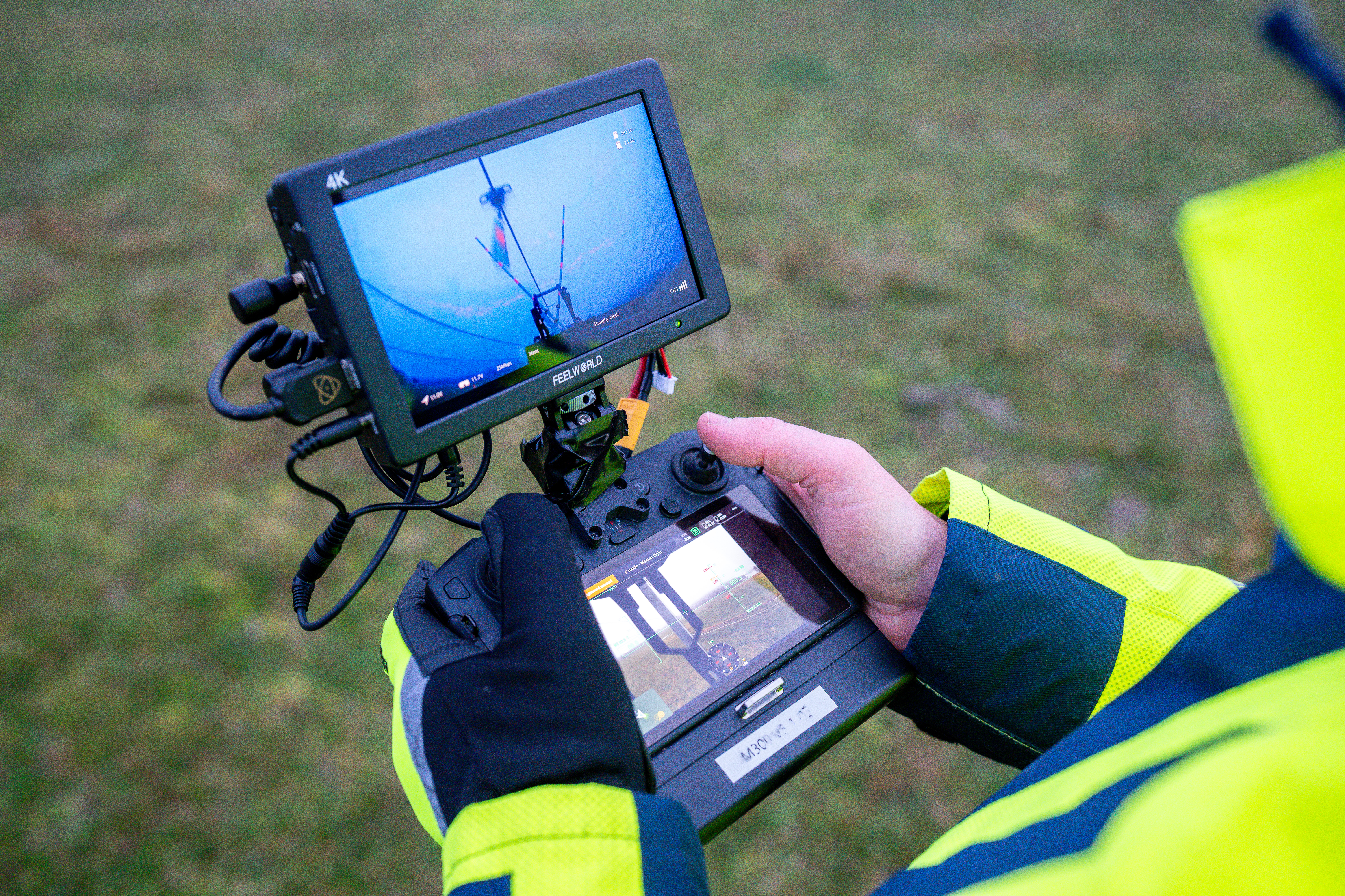 En person med handskar styr en drönare med en fjärrkontroll som visar direktsänd bild över ett gräsområde.