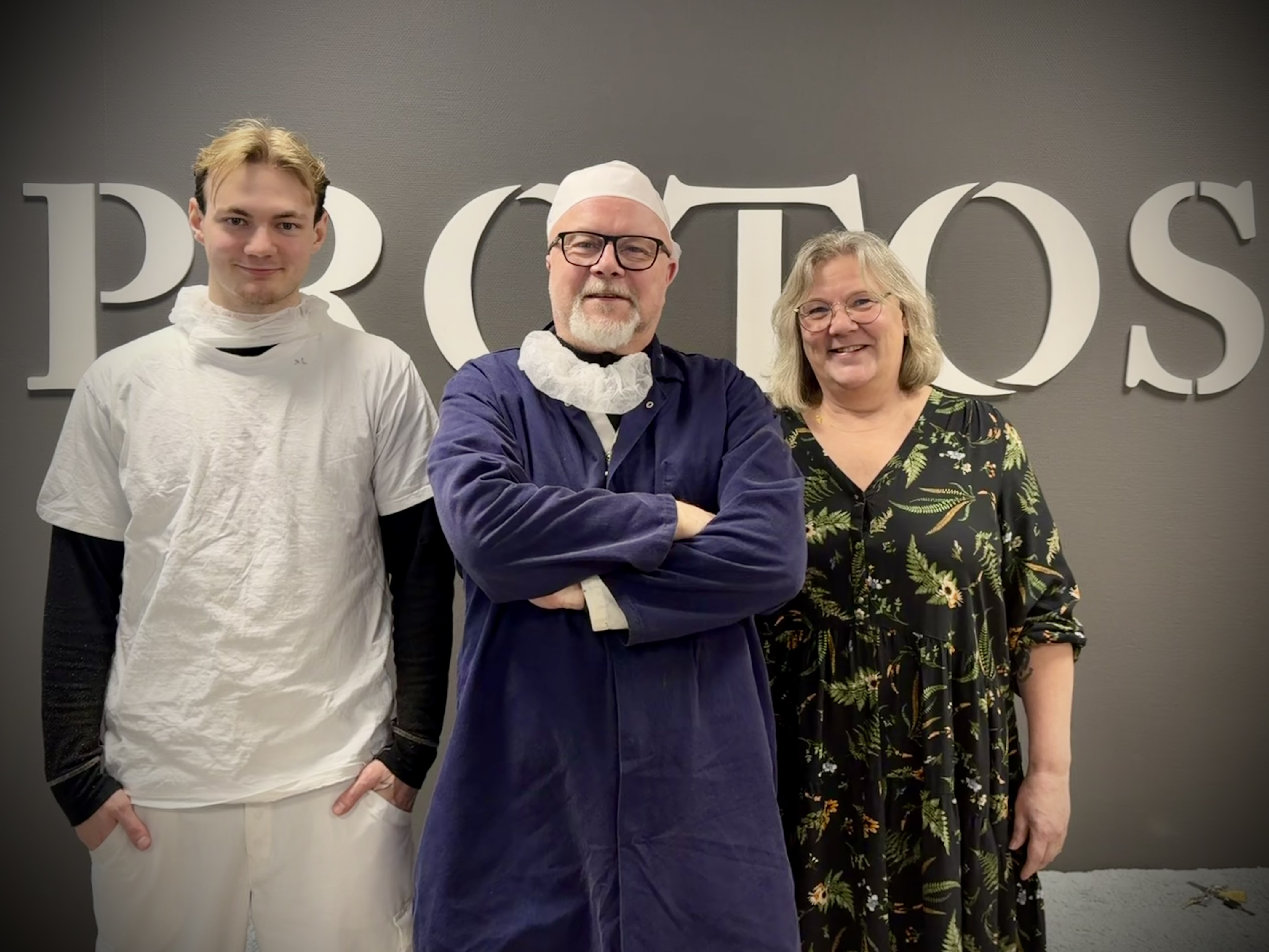 Tre personer står framför en vägg med stora "PROTOS"-bokstäver; en har kockkläder, de andra två har vardags- och blommiga kläder.