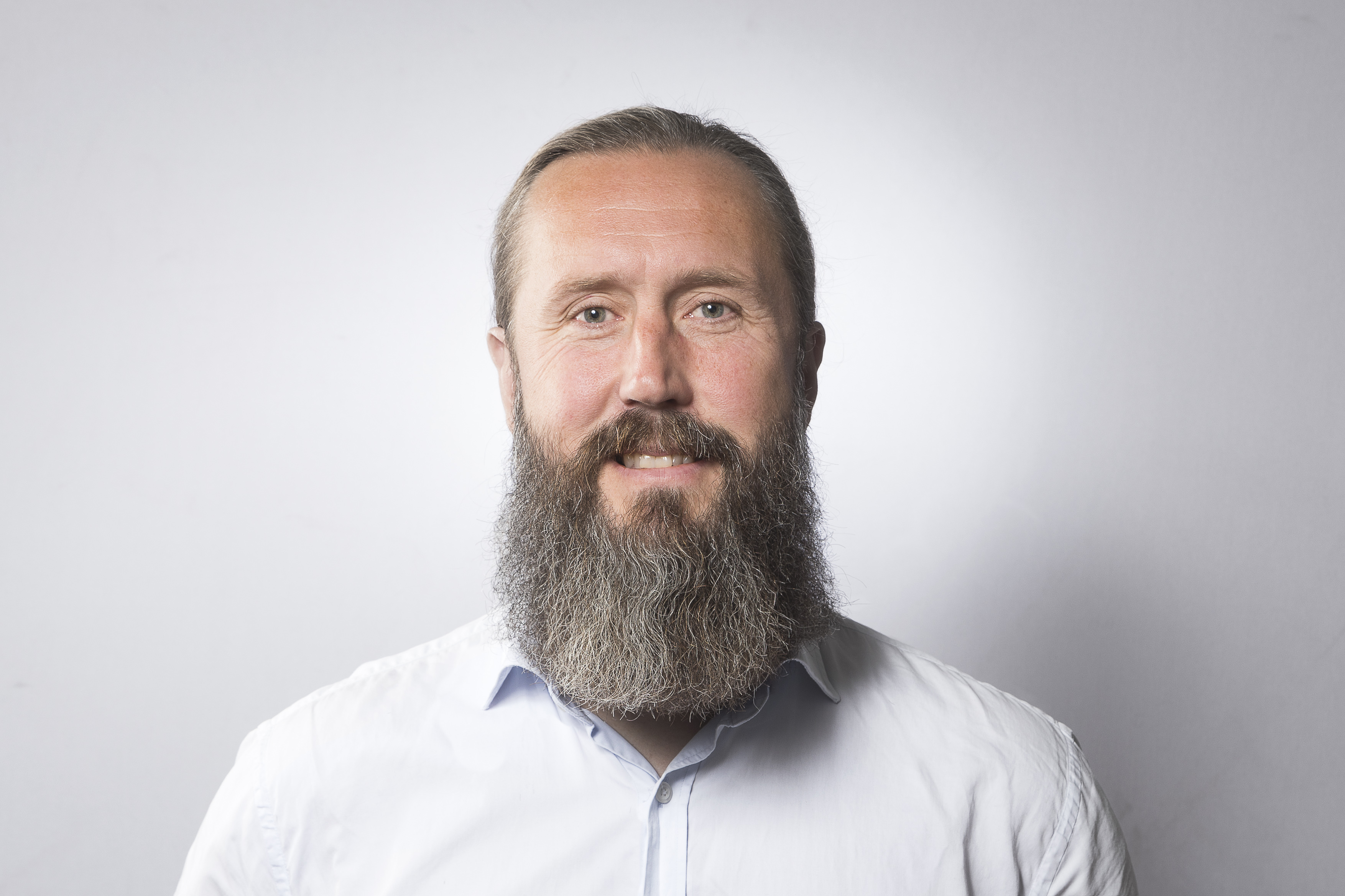 En man med långt skägg och mustasch, har en ljus skjorta på sig och står mot en enfärgad bakgrund och tittar in i kameran.