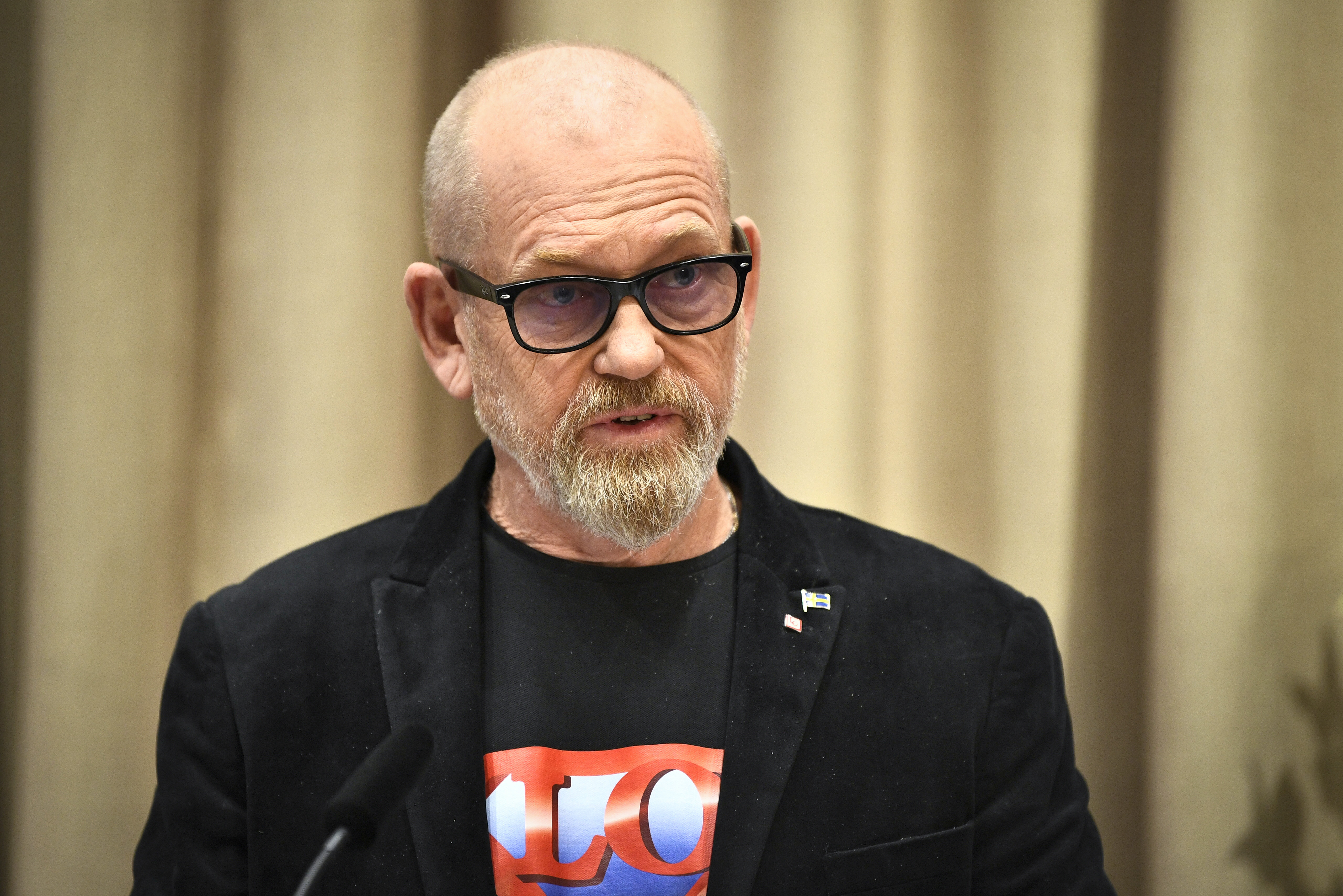 Man i svart kavaj och glasögon står mot en gardin.