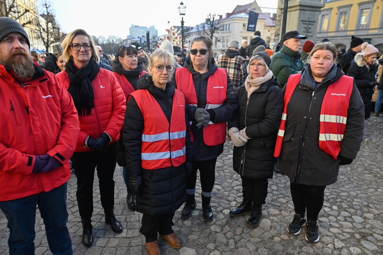 En grupp personer iklädda Kommunals röda västar står tillsammans på en kullerstensbelagd gata, omgivna av många andra människor.