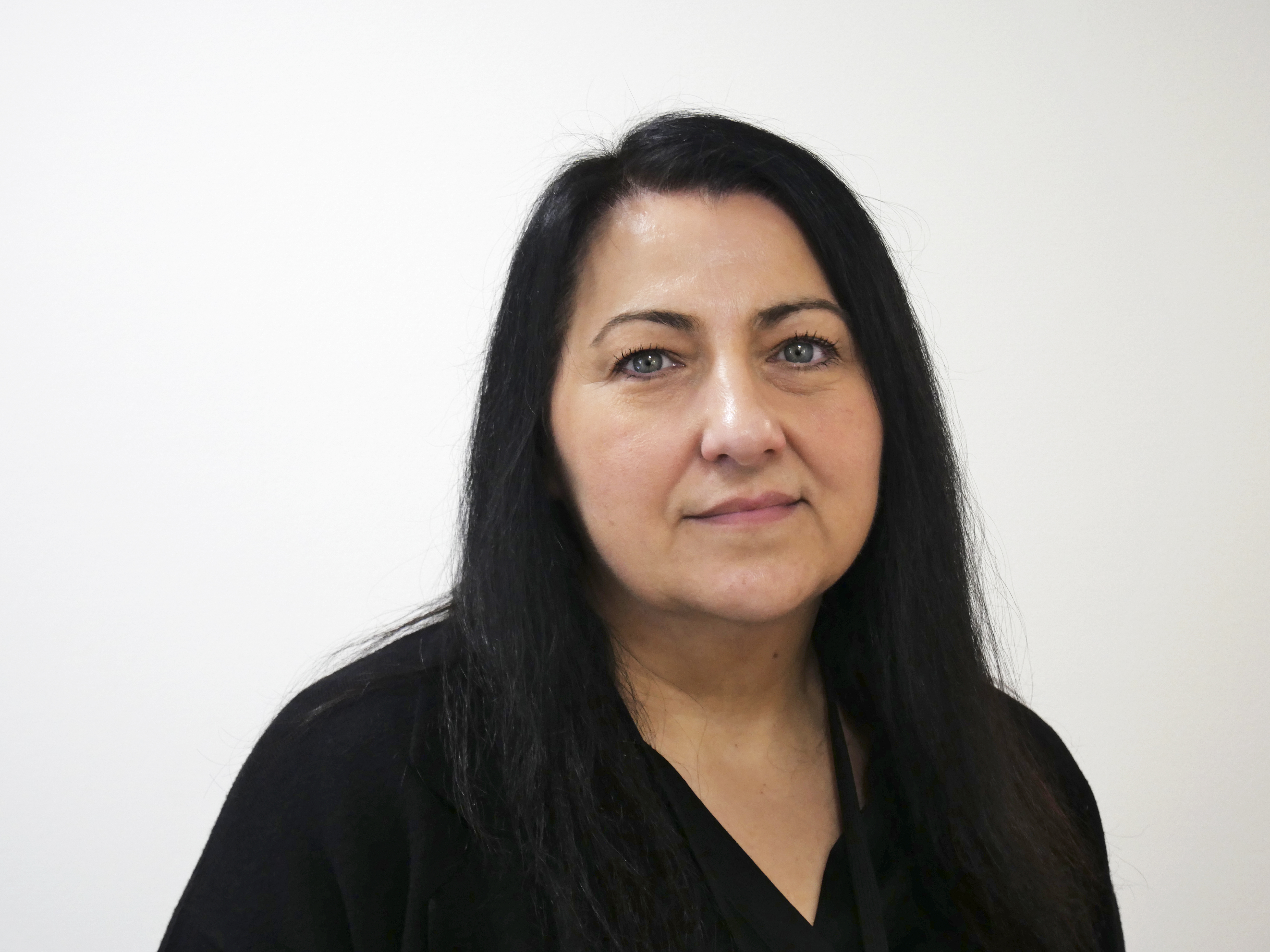 En kvinna med långt svart hår och neutral min står framför en vit bakgrund, iklädd en svart topp.