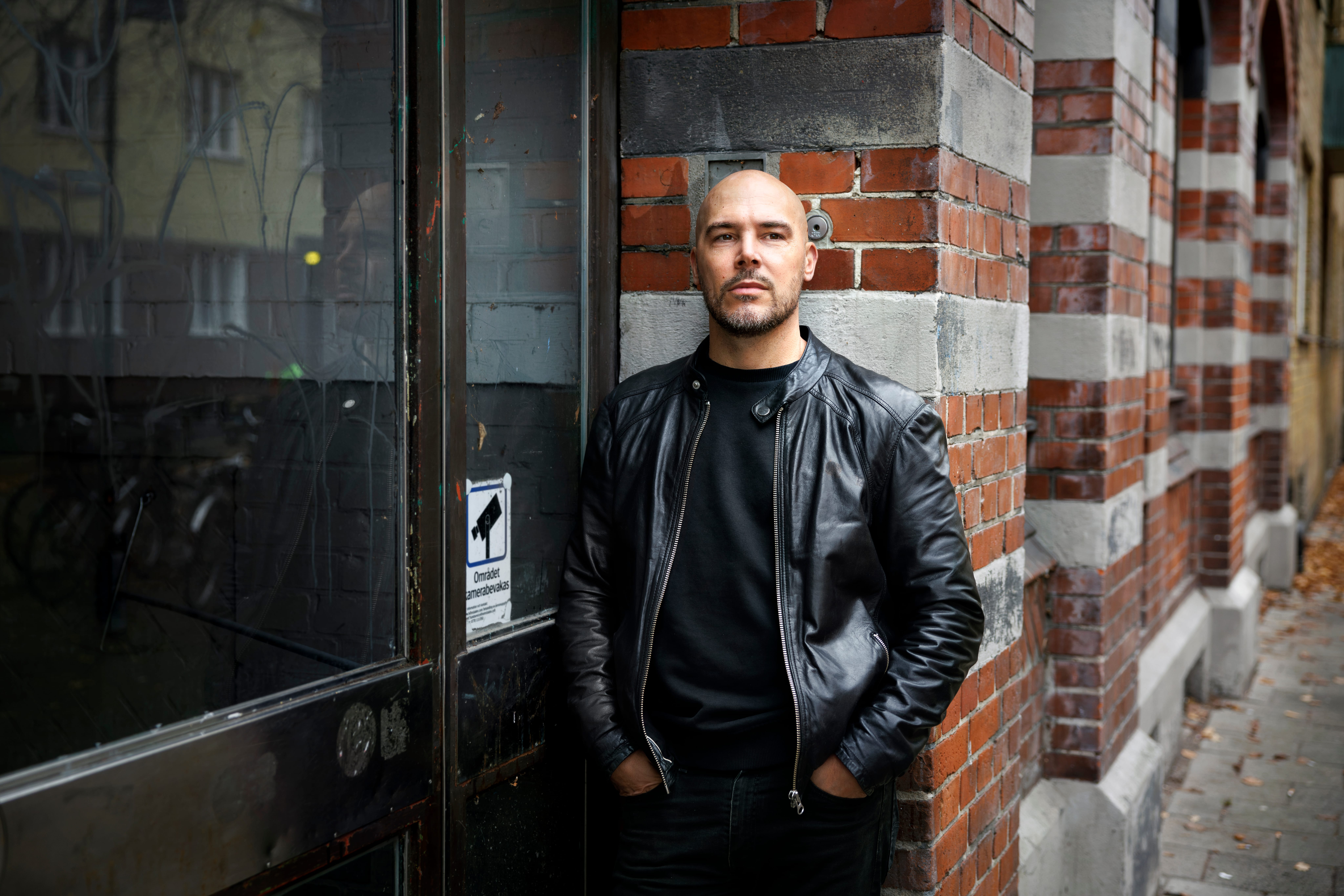 En man i svart skinnjacka står mot en tegelvägg bredvid en glasdörr med en liten säkerhetsdekal.