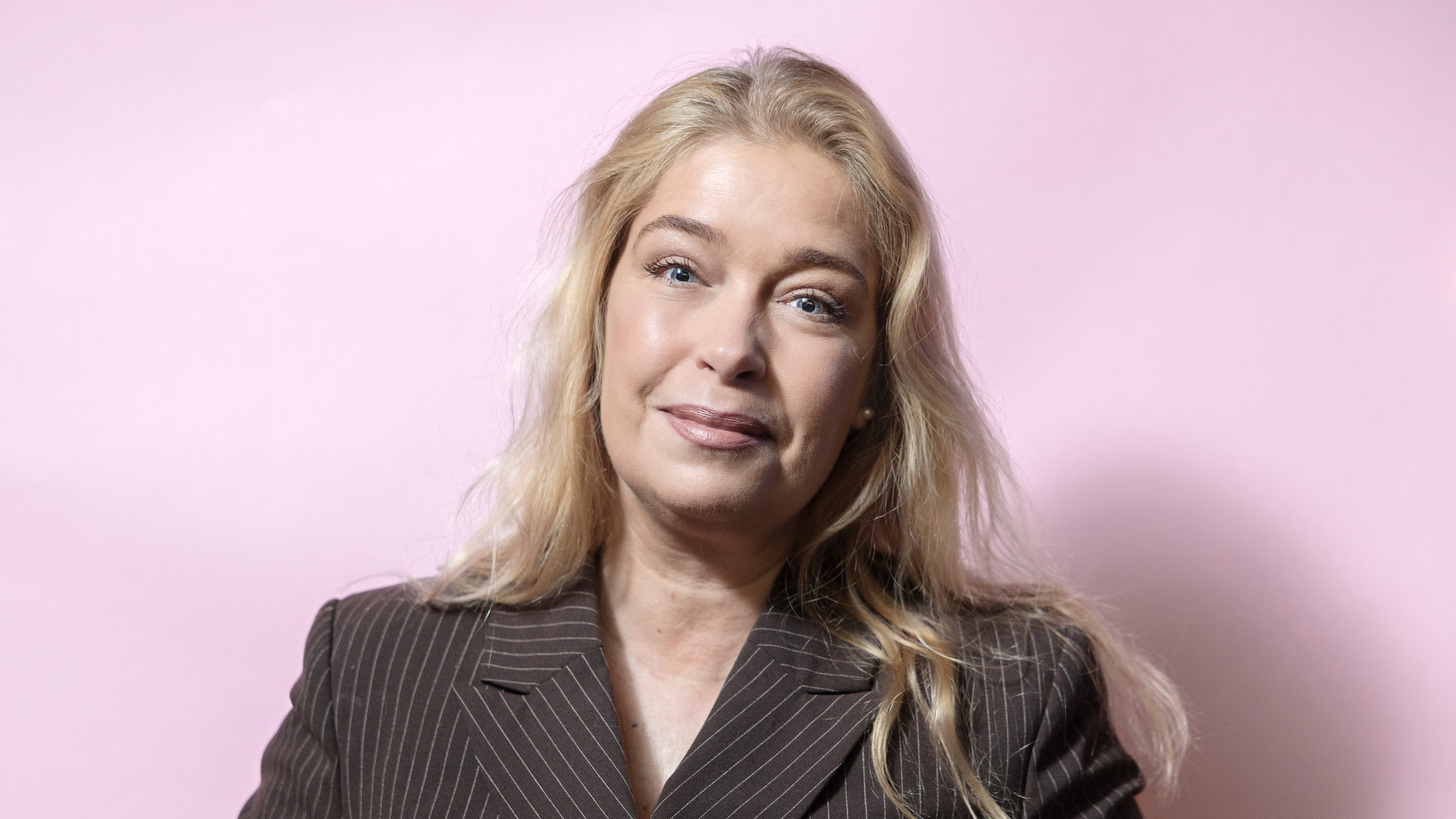 En kvinna med långt blont hår och en brunrandig kavaj står framför en rosa bakgrund.