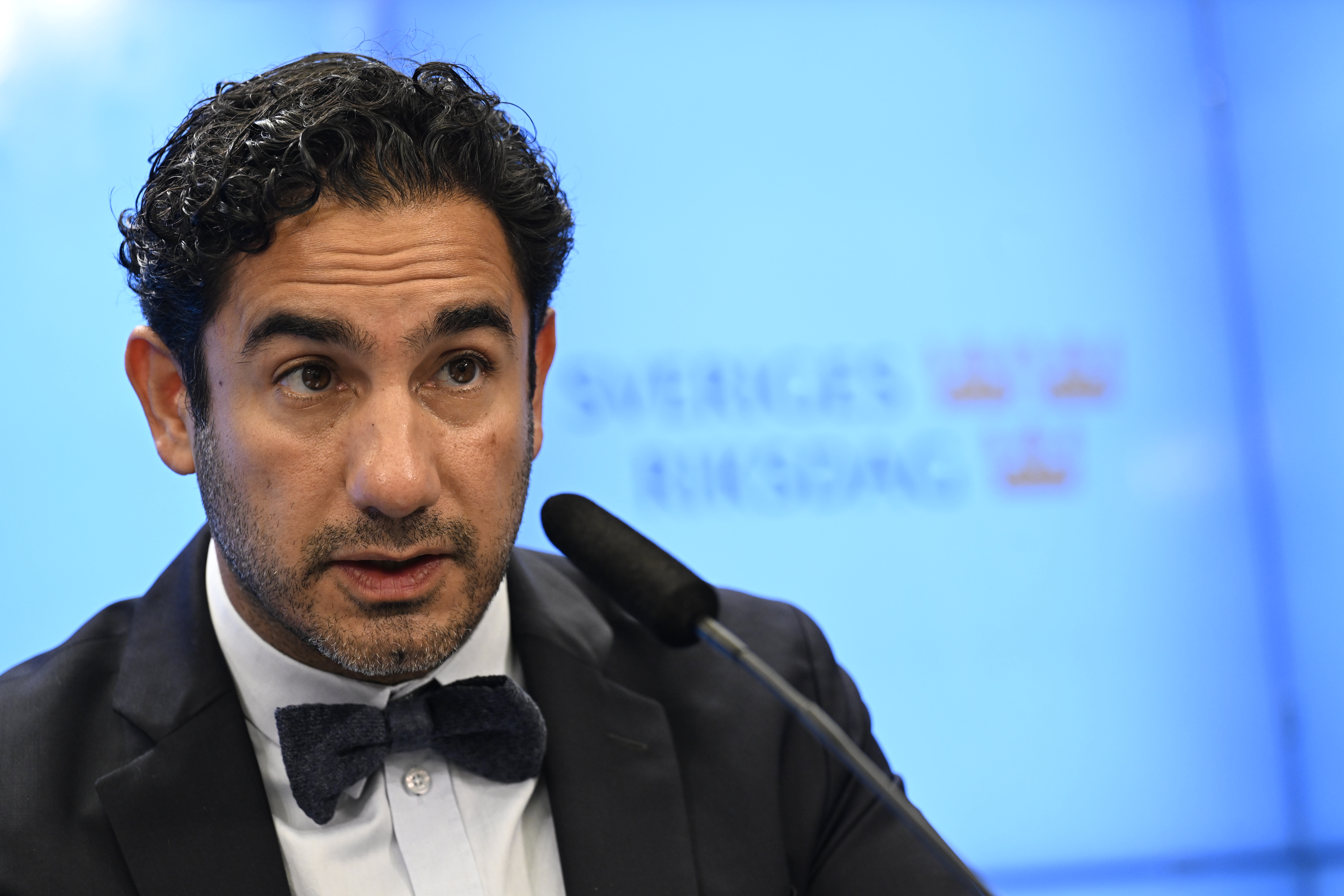 En man i kostym och fluga talar vid en mikrofon framför en blå bakgrund med texten "Sveriges Riksdag".